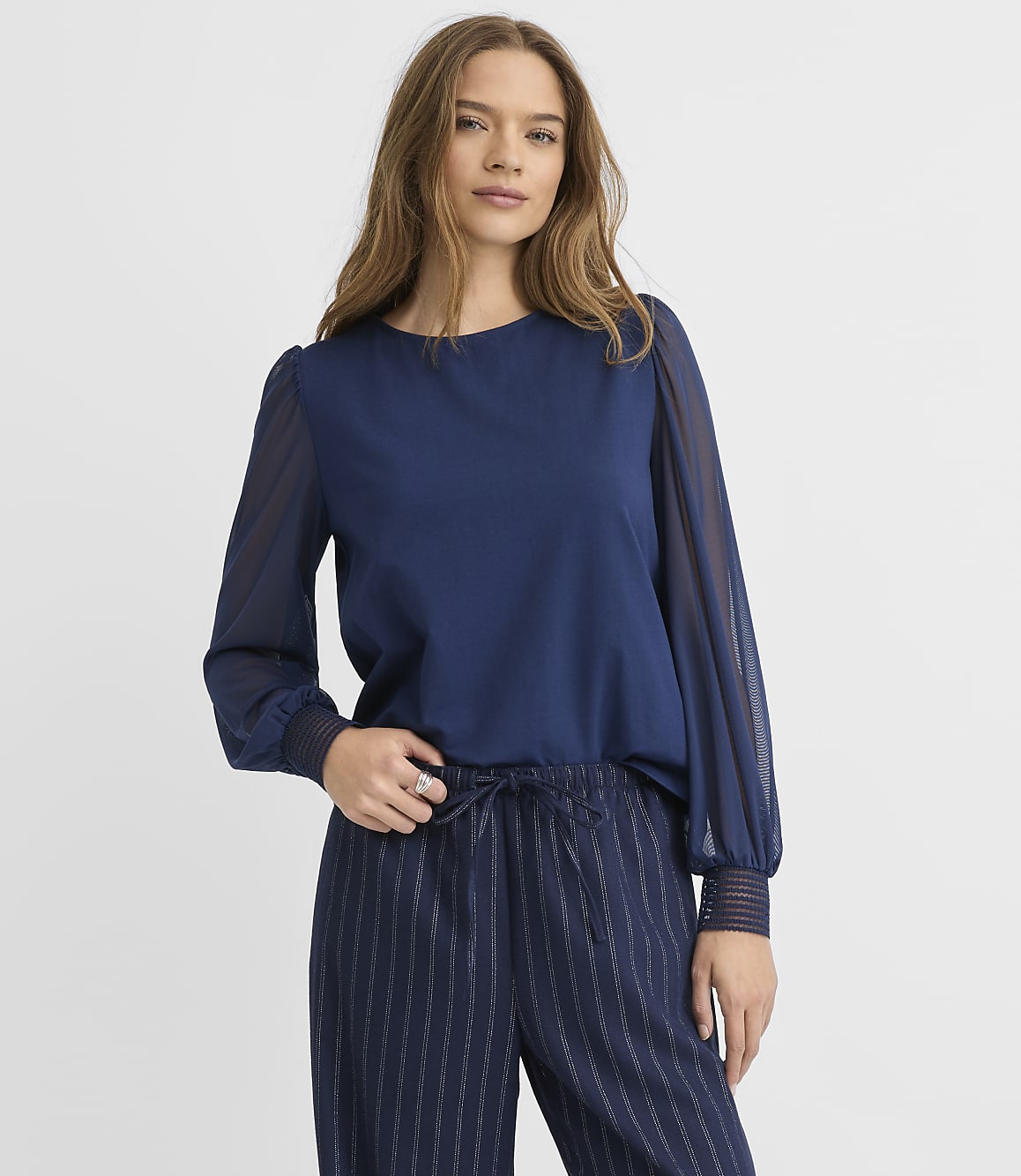 A woman wearing a navy blue blouse and pinstriped trousers stands against a plain white background.