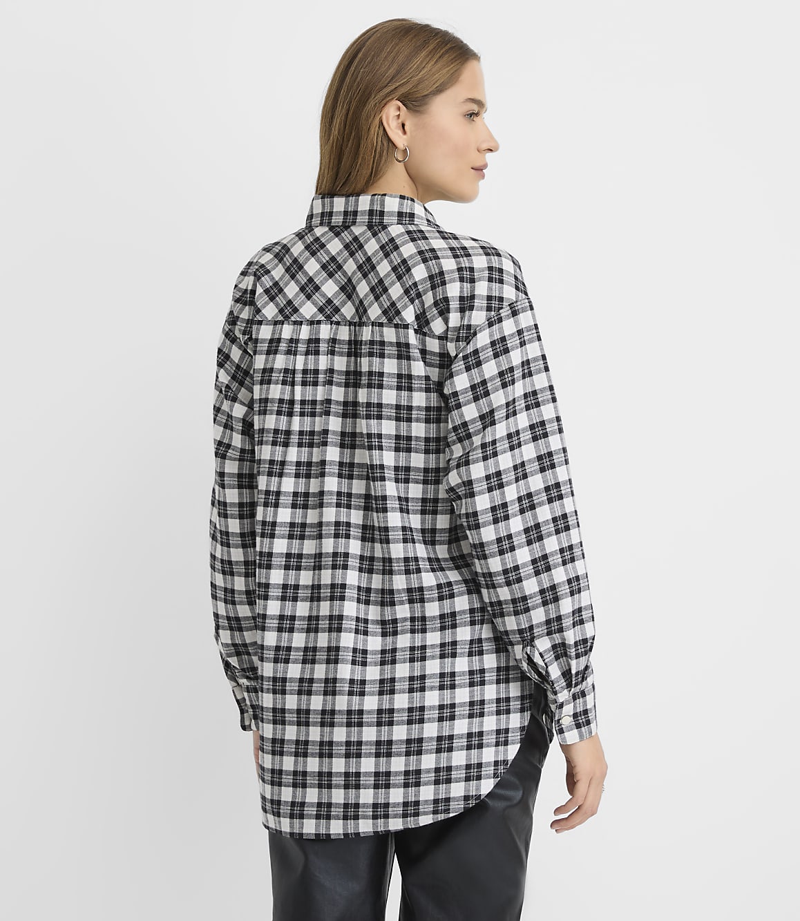 A woman wearing a black and white plaid shirt and black leather pants stands against a plain white background.