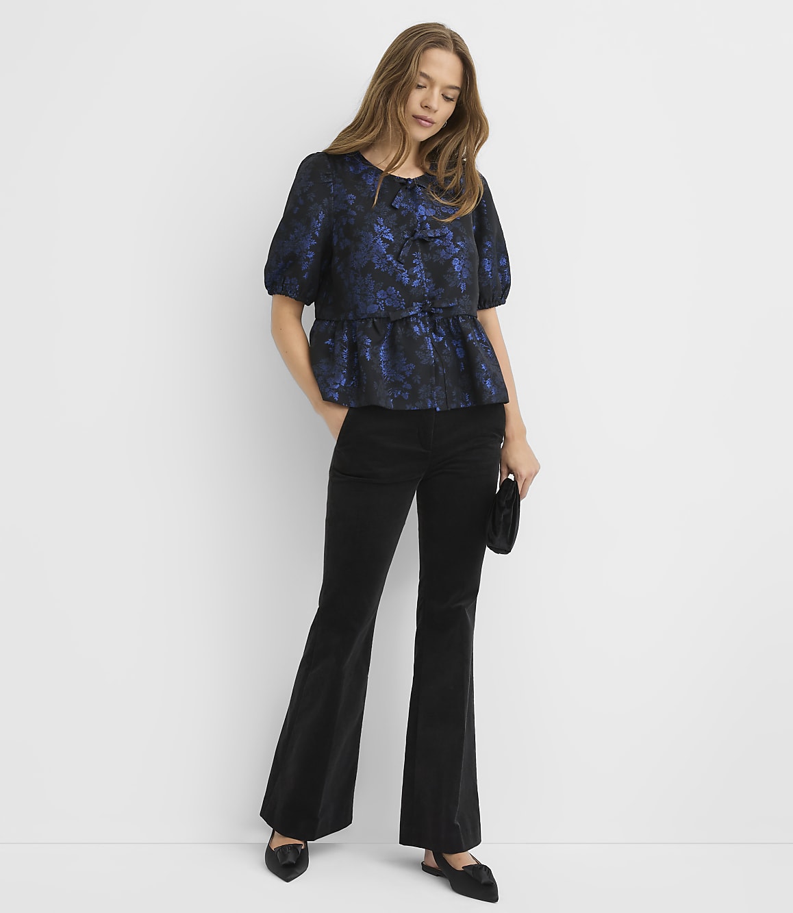 A woman wearing a blue floral blouse and black flared trousers stands against a plain white background.