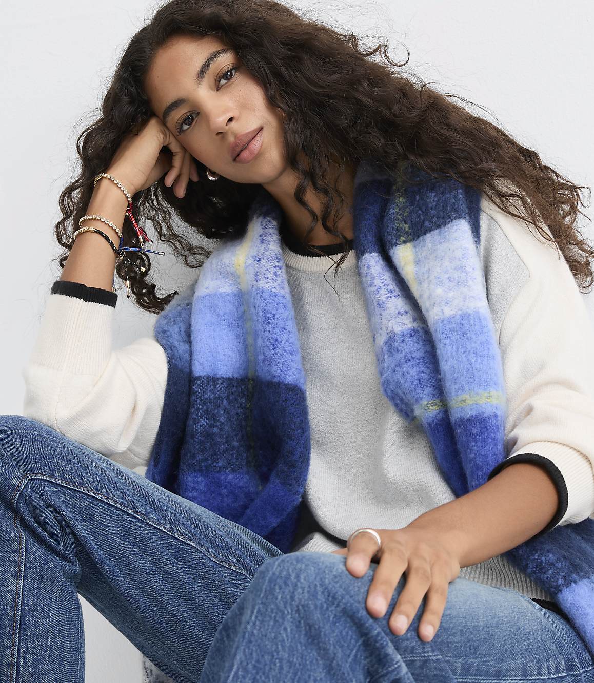 A woman with long curly hair wearing a white sweater, blue jeans, and a blue plaid scarf poses against a plain light gray background.