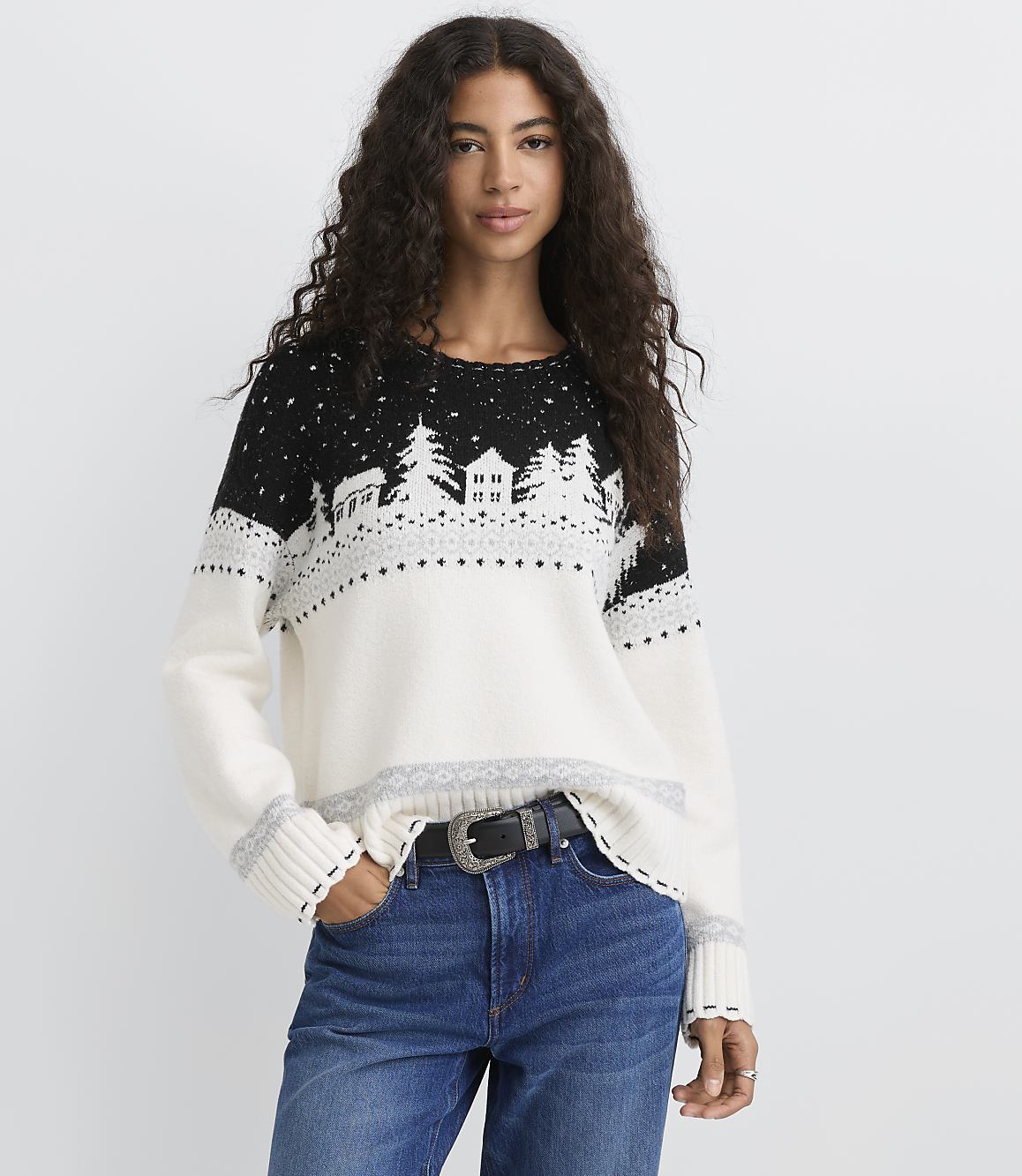 A woman with long curly hair wearing a black and white winter-themed sweater and blue jeans stands against a plain white background.