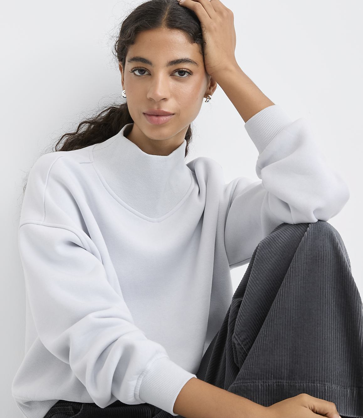 A woman wearing a light blue sweatshirt and dark grey corduroy pants poses against a plain white background.