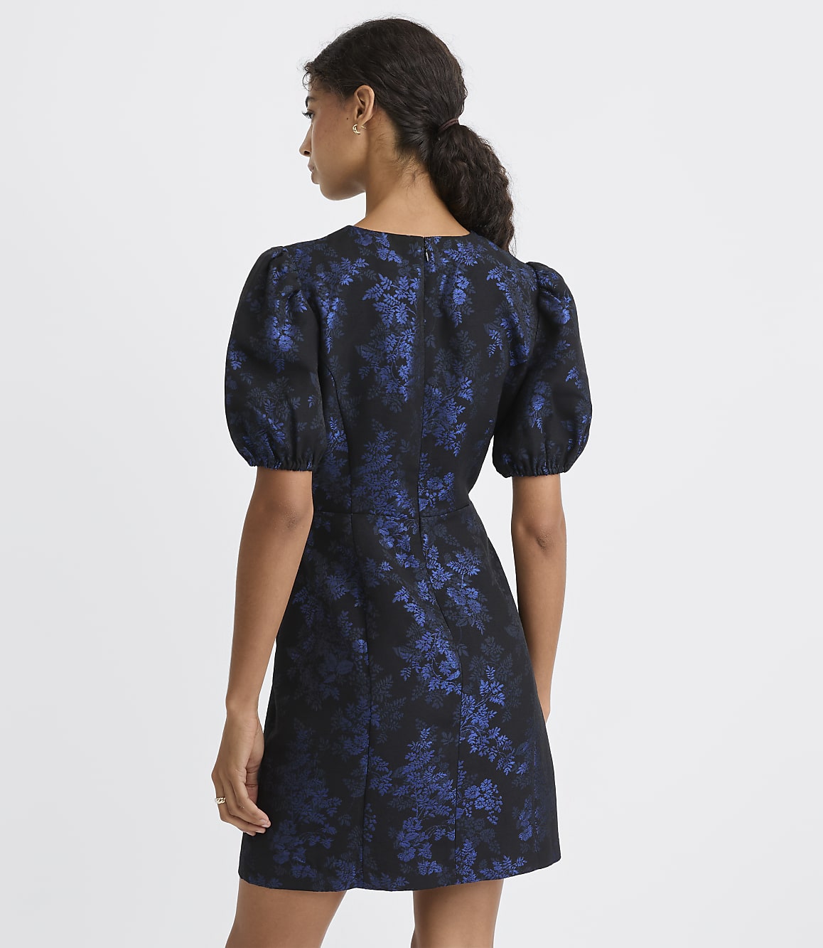 A woman stands with her back to the camera wearing a short, puff-sleeved blue and black floral dress against a plain white background.