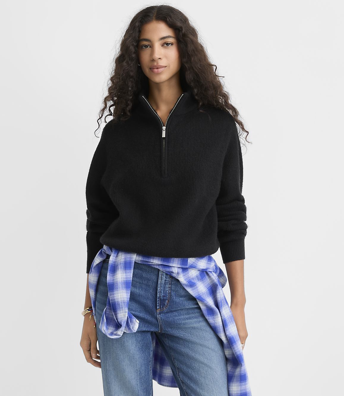 A woman wearing a black quarter-zip sweater and blue jeans with a plaid shirt tied around her waist stands against a plain white background.