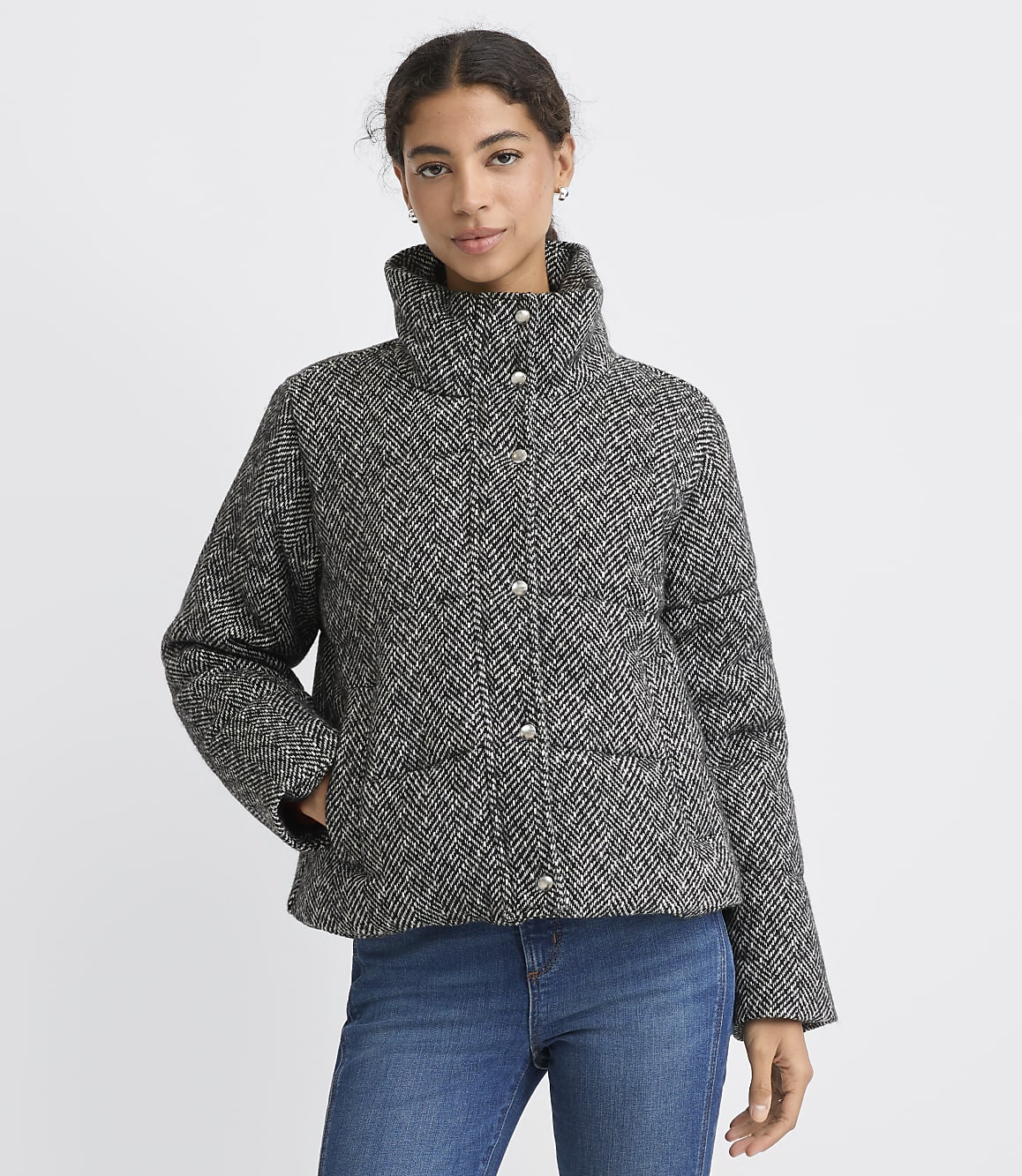 A woman wearing a grey and black herringbone patterned jacket and blue jeans stands against a plain white background.