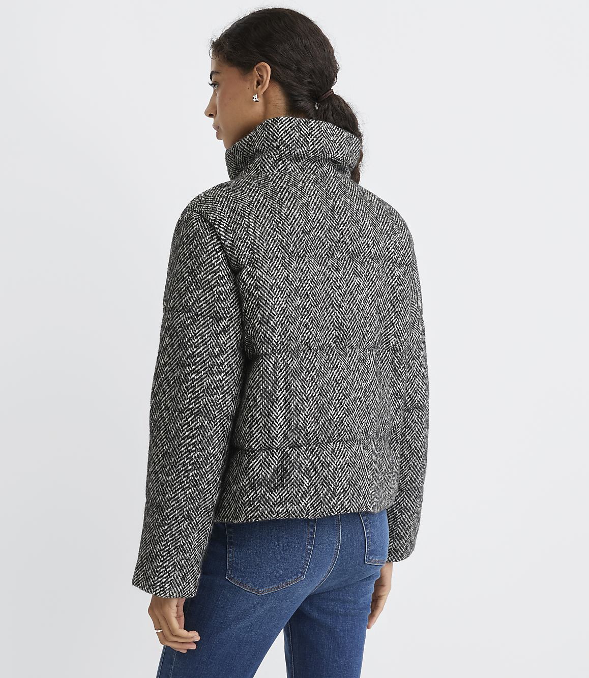 A woman wearing a black and white herringbone puffer jacket and blue jeans stands against a plain white background.