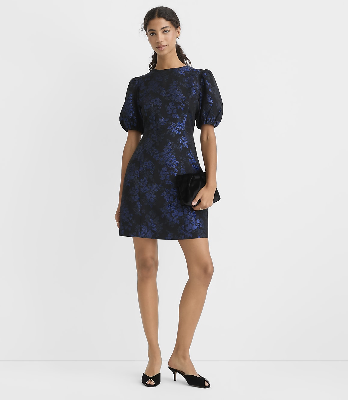 A woman wearing a navy blue floral puff-sleeve dress and black heels stands against a plain white background.