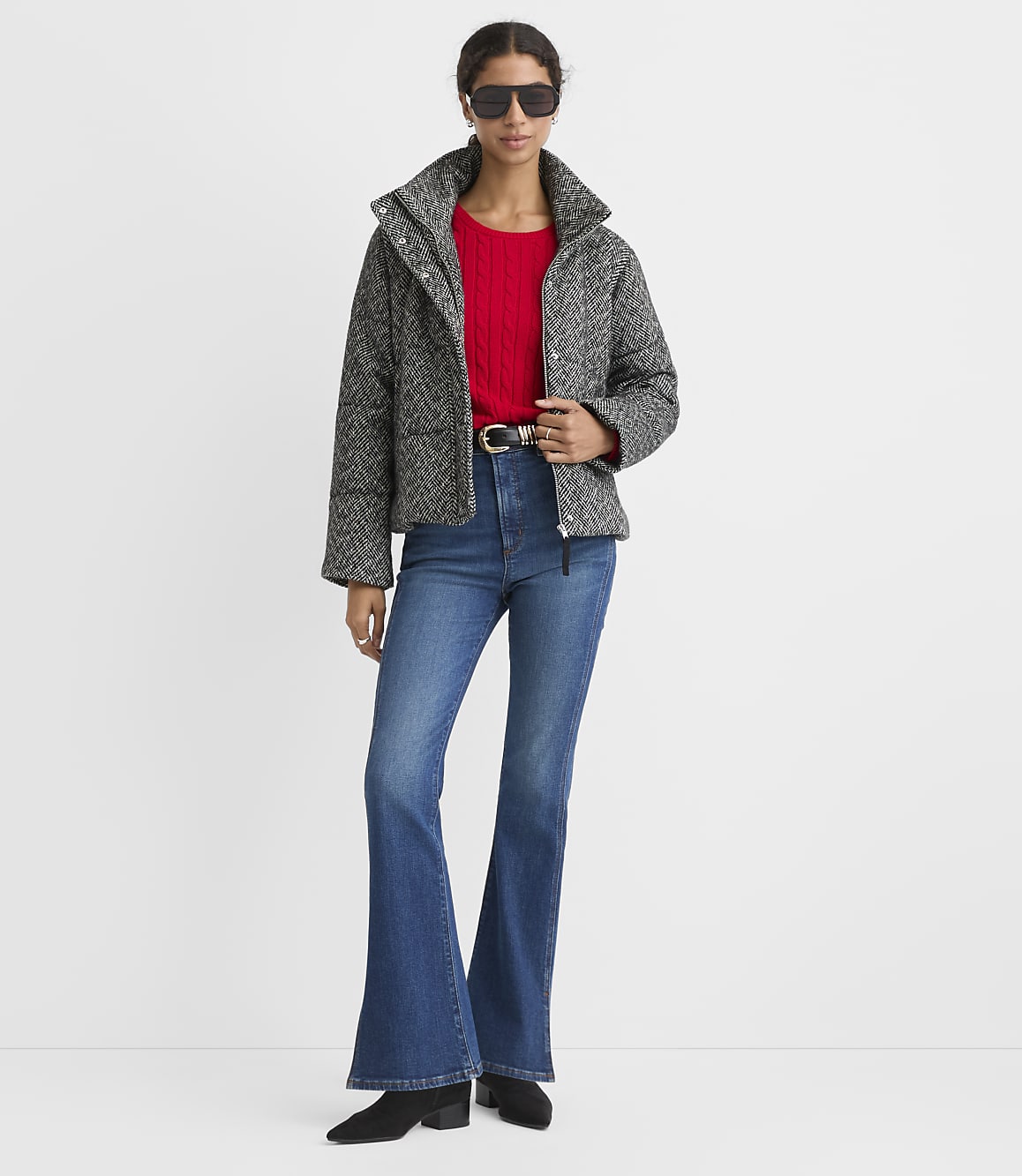 A woman wearing a grey patterned jacket, red sweater, and flared blue jeans stands against a plain white background.