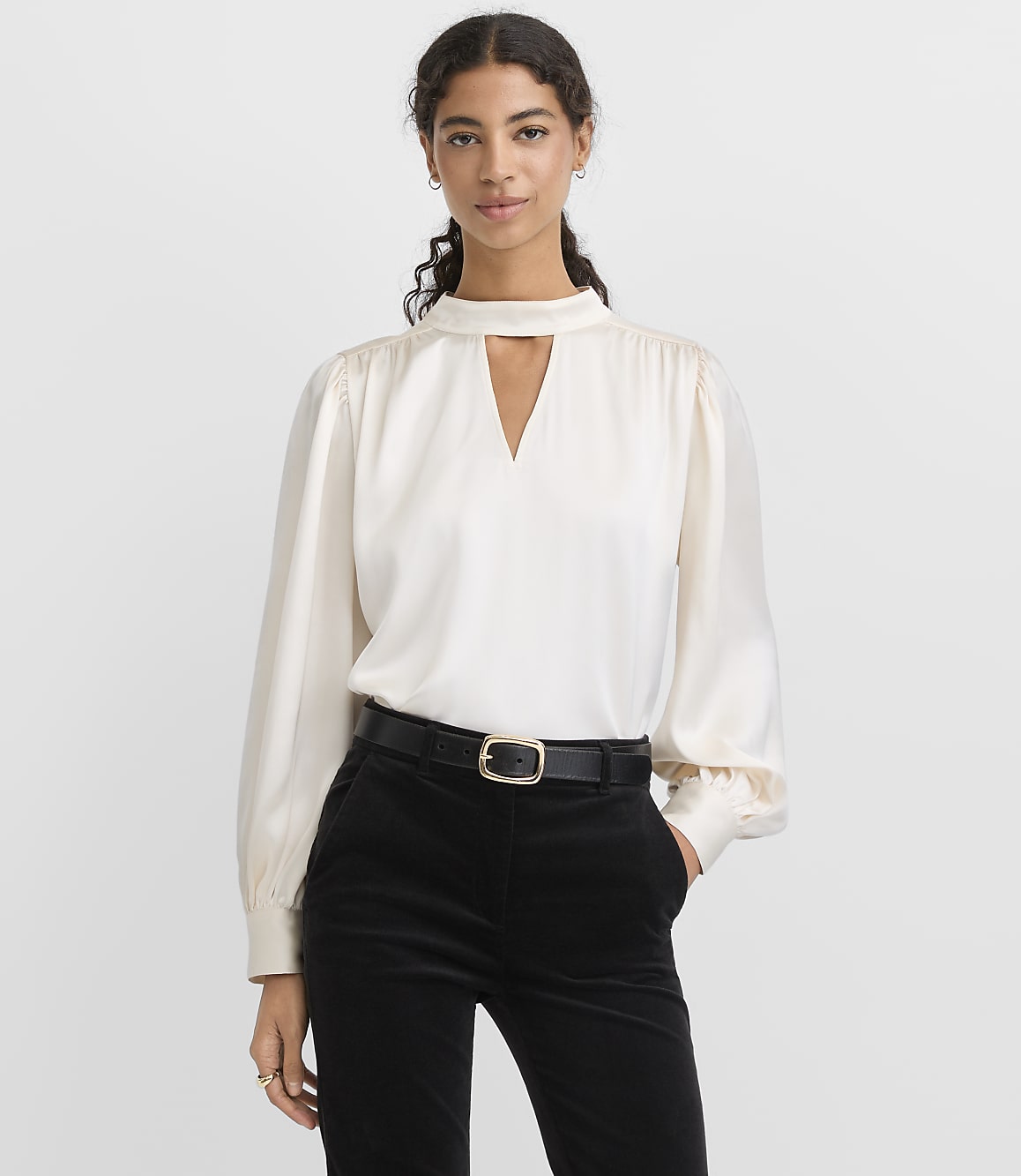 A woman wearing a white long-sleeved blouse and black trousers stands against a plain light gray background.