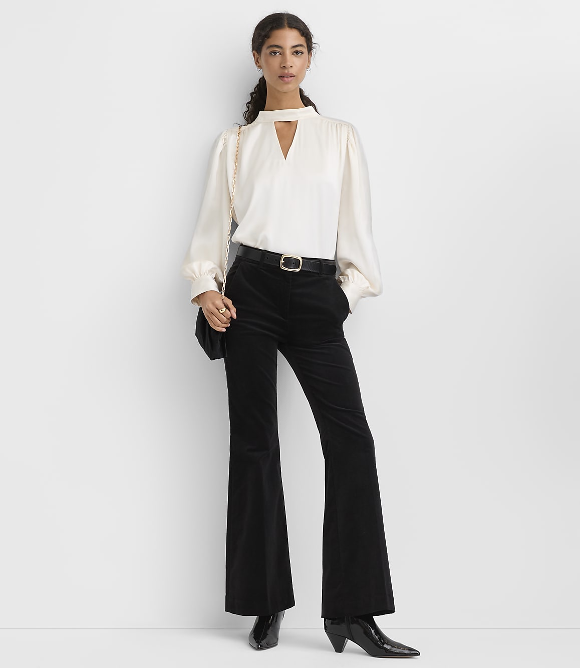 A woman wearing a white blouse and black flared trousers stands against a plain white background.