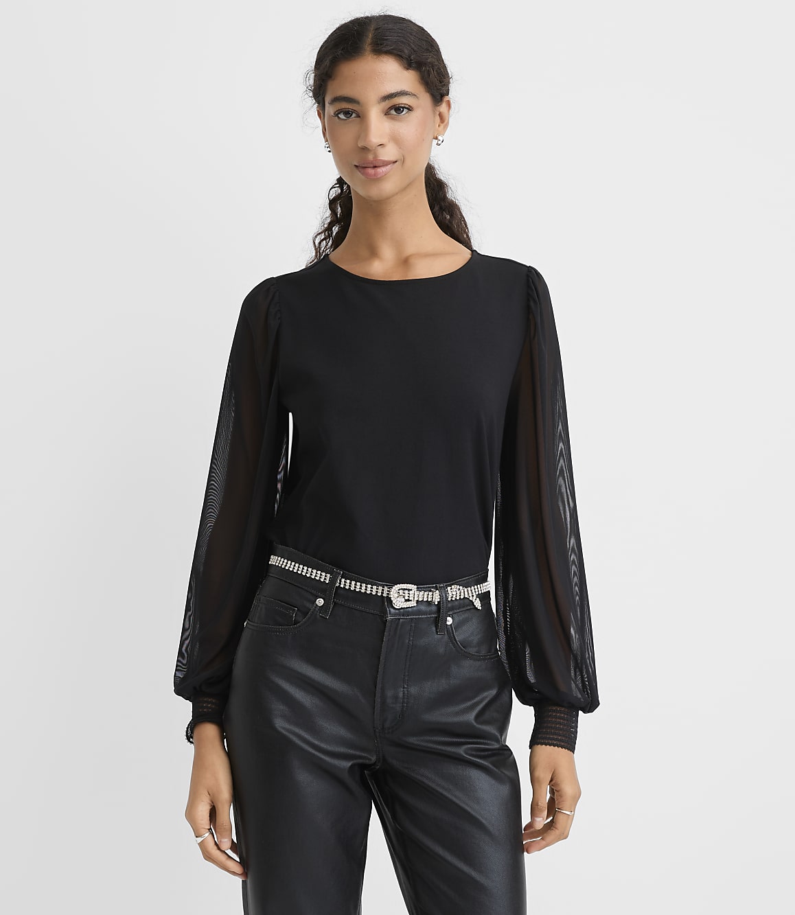 A woman wearing a black top with sheer sleeves and black faux-leather pants stands against a plain white background.