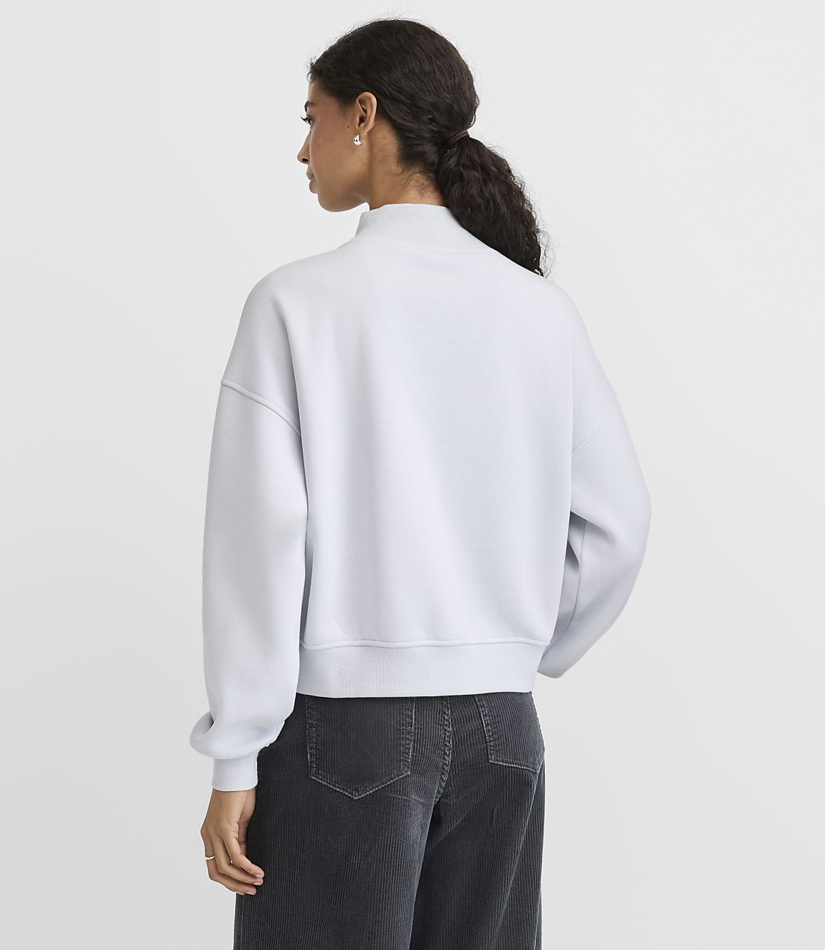 A woman wearing a light blue sweatshirt and dark corduroy pants stands with her back to the camera against a plain white background.