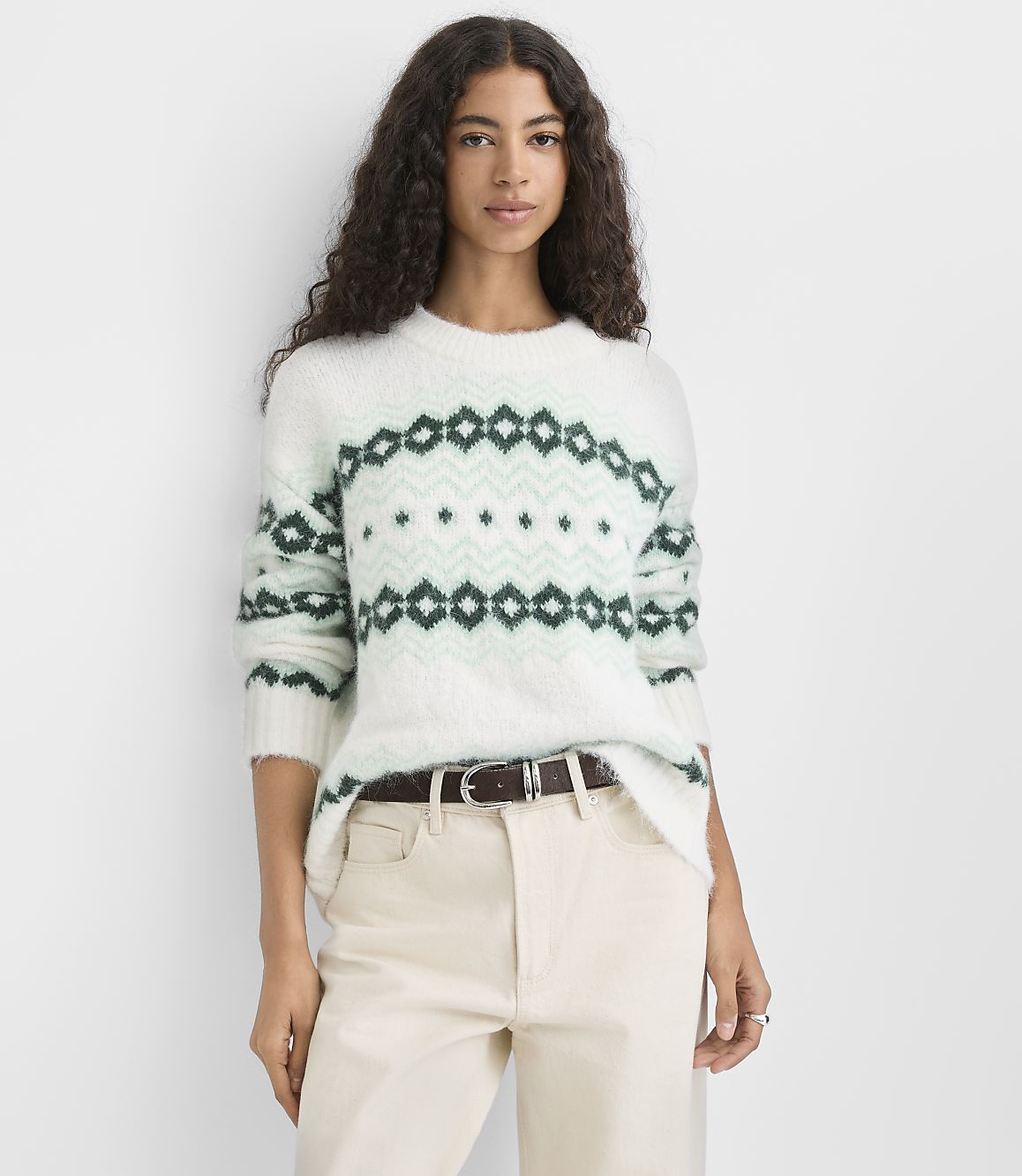 A woman wearing a white and green patterned fuzzy sweater and cream-colored pants stands against a plain white background.