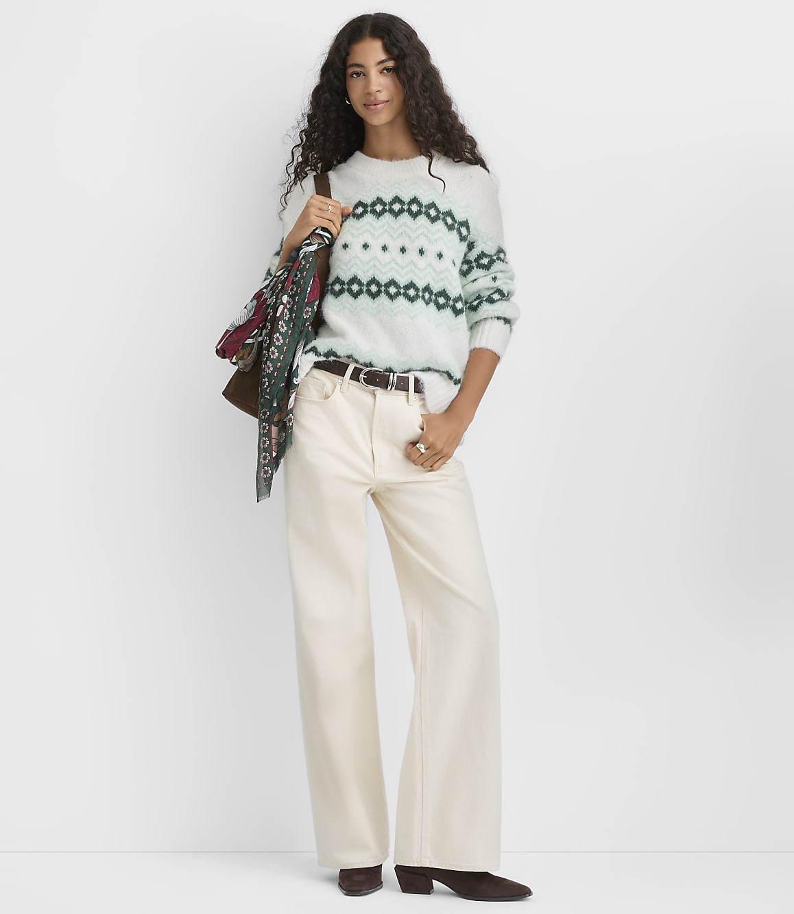 A woman wearing a patterned white and green sweater and wide-leg cream pants stands against a plain white background.