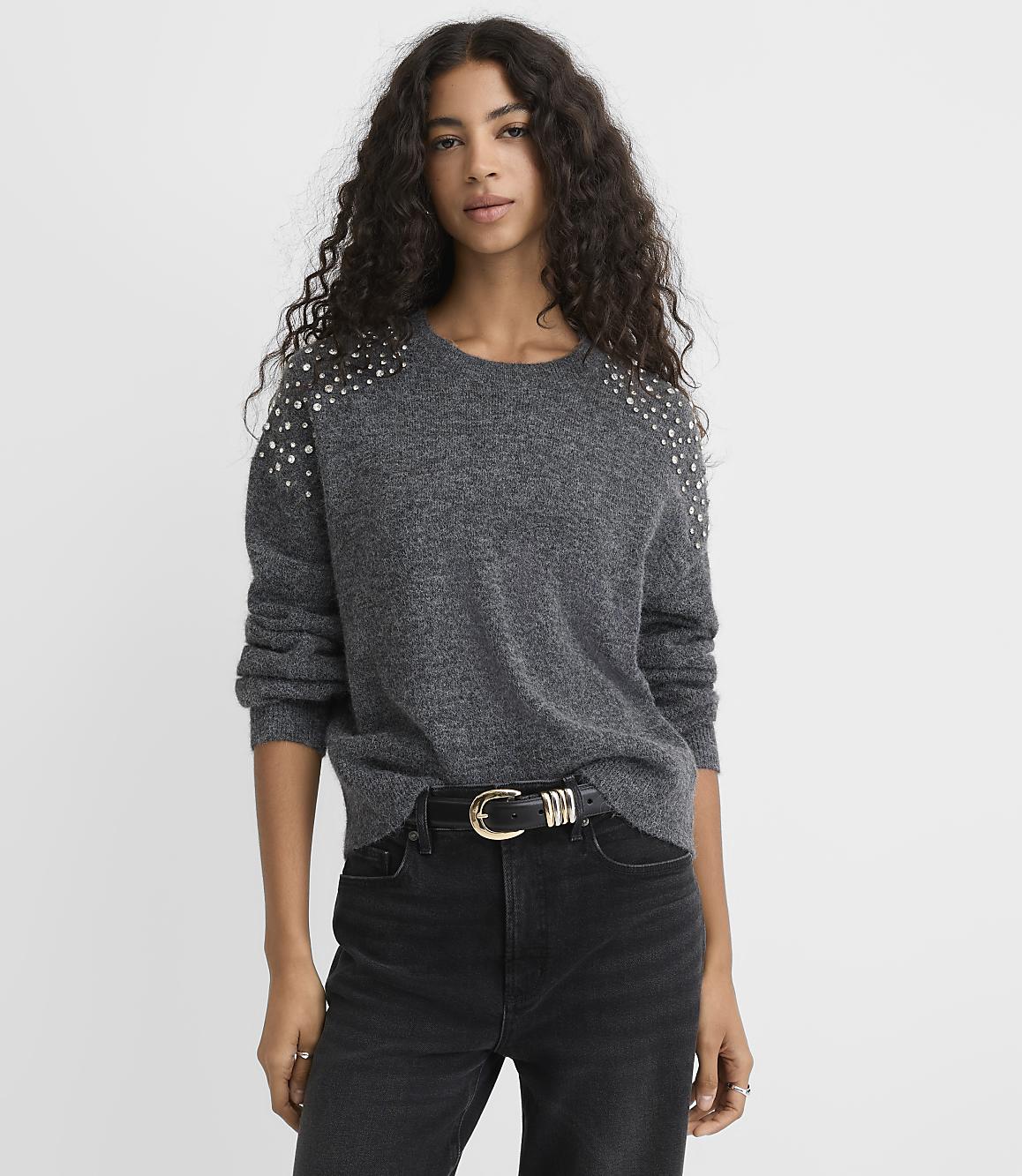 A woman with long curly hair wears a grey studded sweater and black jeans against a plain white background.