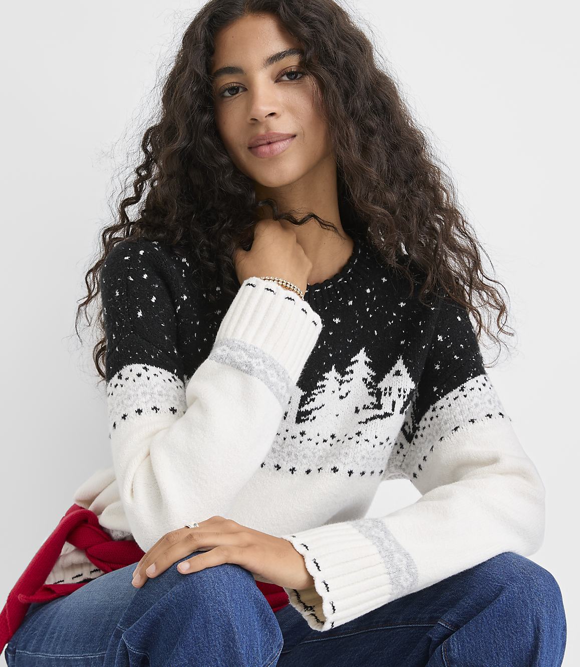 A woman with long curly hair wearing a black and white winter-themed sweater and blue jeans poses against a plain white background.