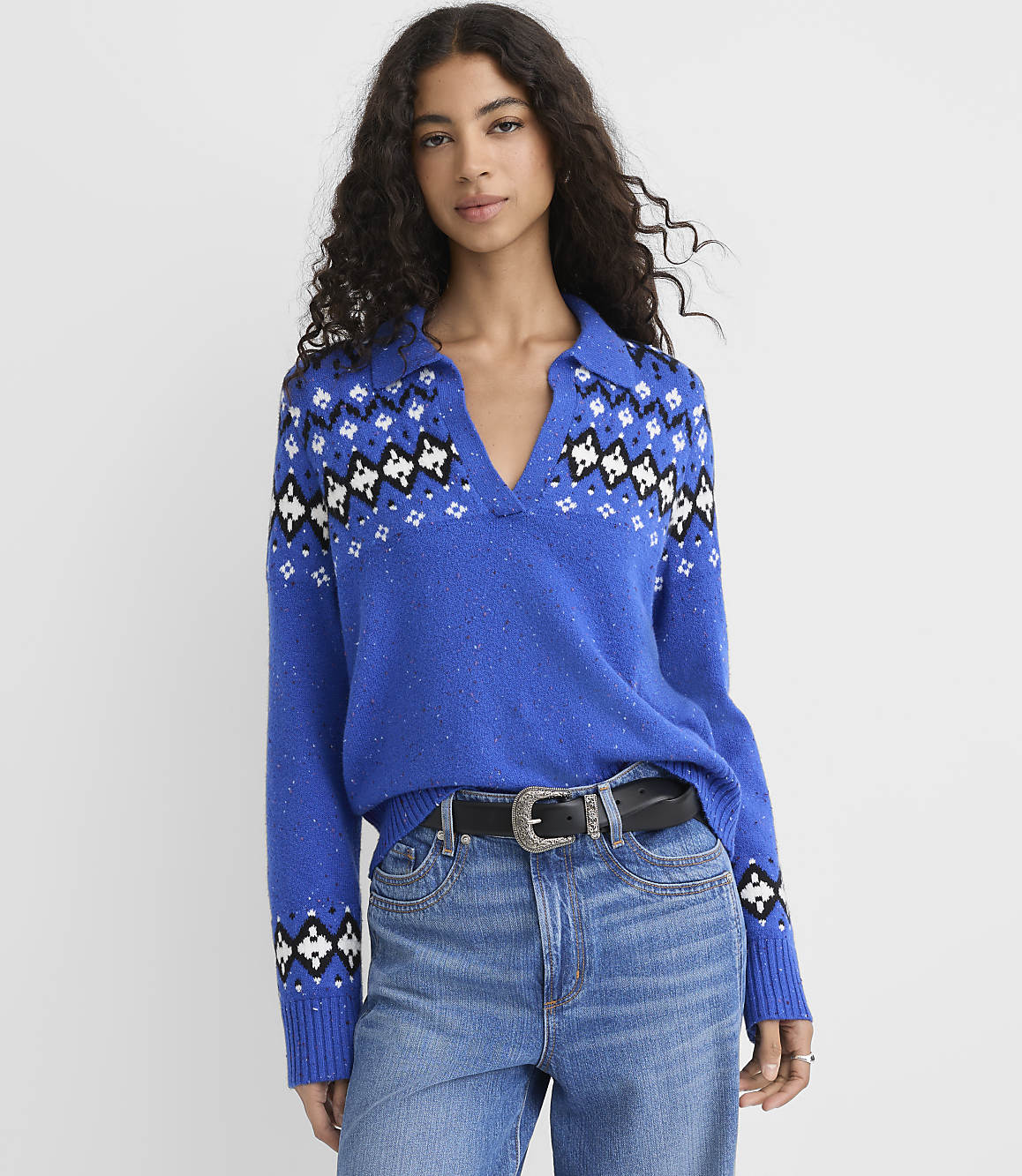 A woman wearing a blue patterned sweater and jeans stands against a plain white background.