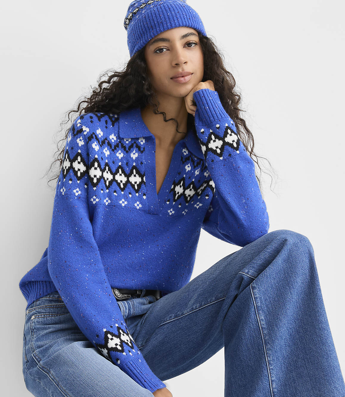 A woman wearing a blue patterned sweater, blue beanie, and jeans poses against a plain white background.