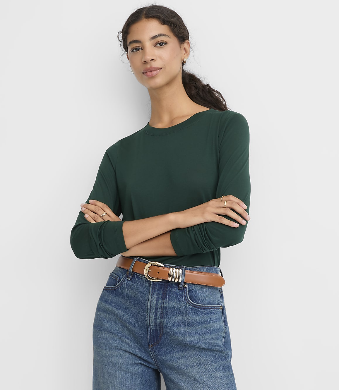 A woman wearing a dark green long-sleeved shirt and blue jeans with a brown belt stands with her arms crossed against a plain white background.