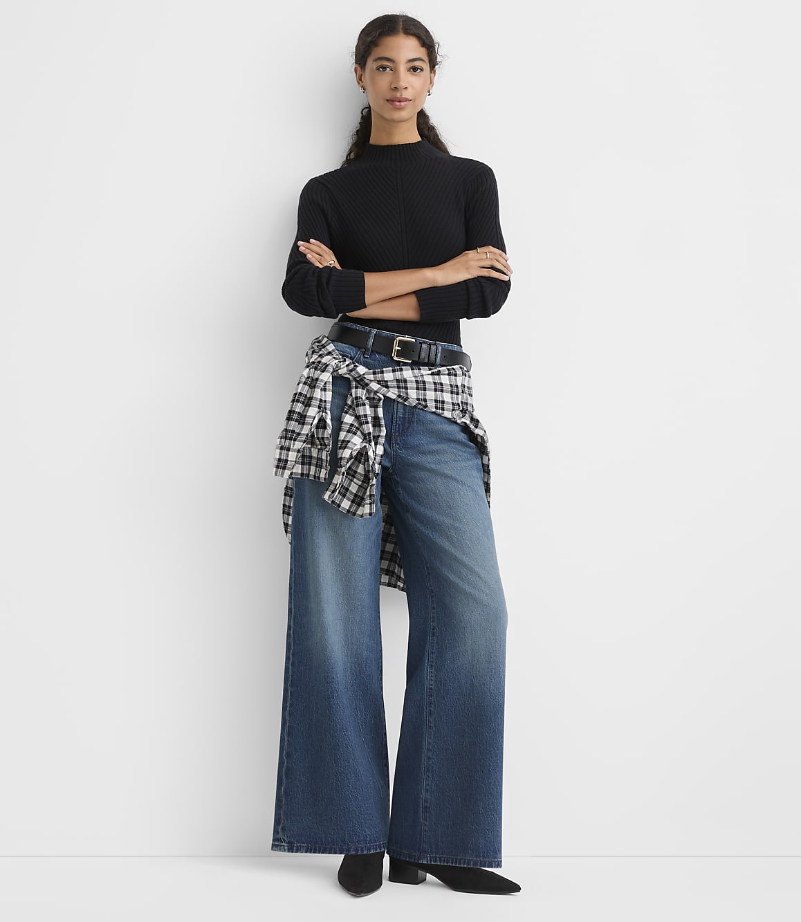 A woman wearing a black ribbed top, wide-leg jeans, and a checkered shirt tied around her waist stands against a plain white background.