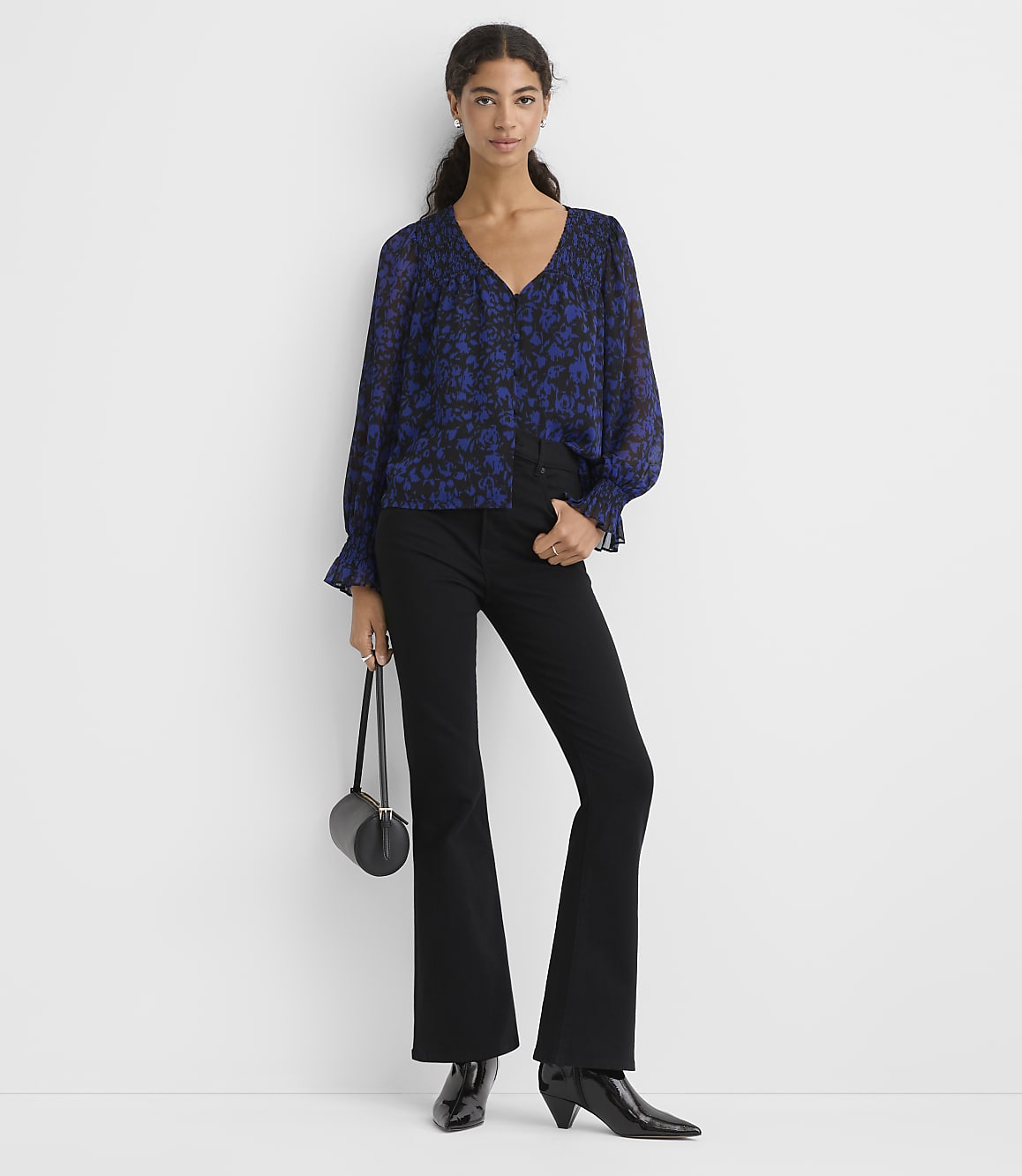 A woman wearing a blue floral blouse and black flared trousers stands against a plain white background.