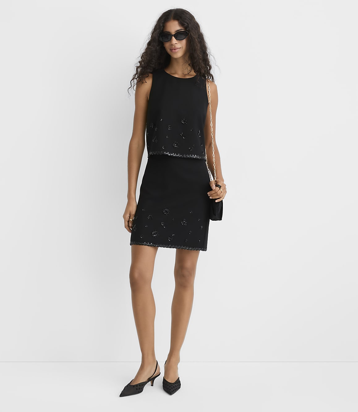 A woman wearing a black sleeveless dress, sunglasses, and black heels stands against a plain white background.