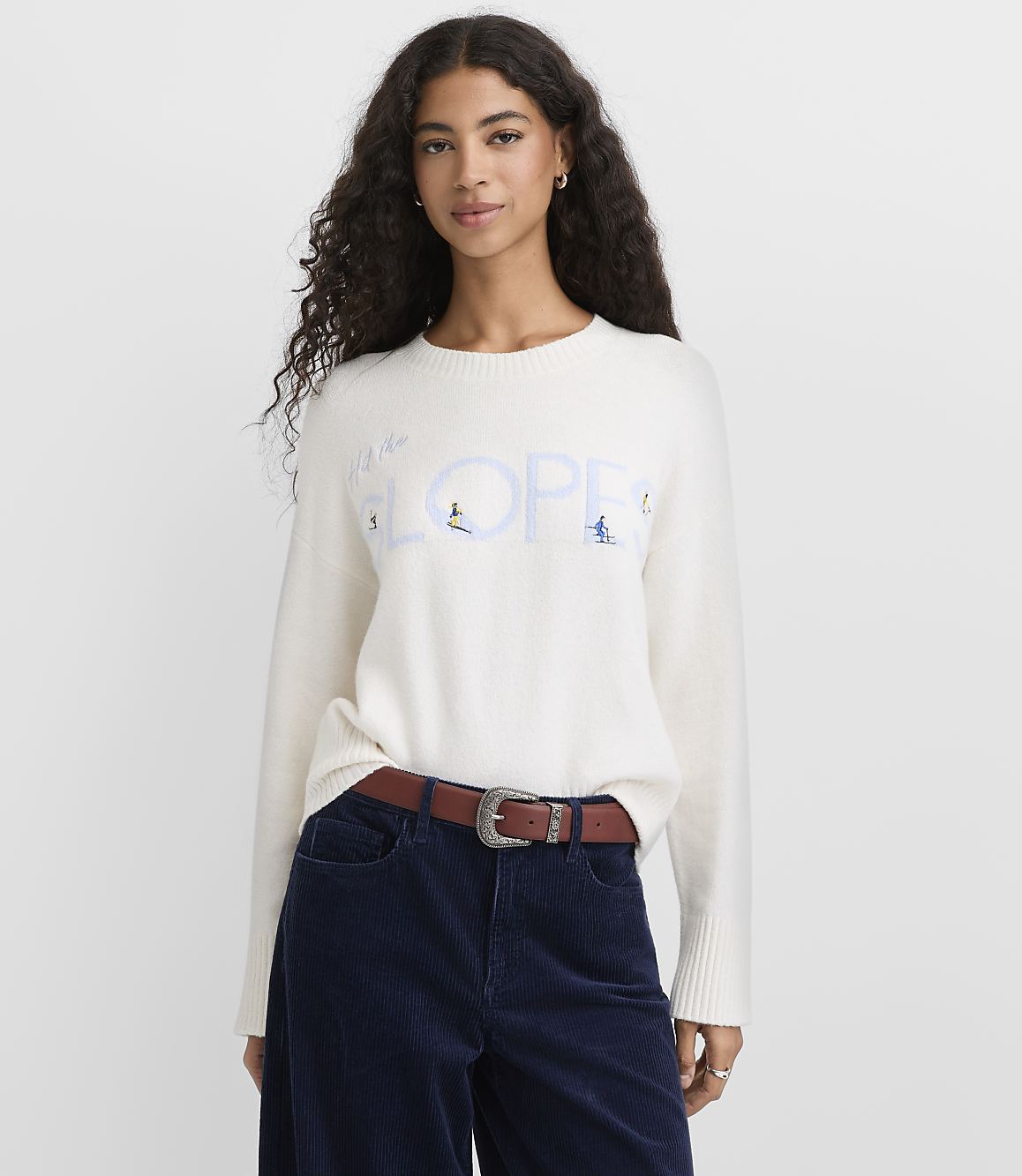 A woman with long curly hair wearing a white graphic sweater and navy corduroy pants stands against a plain light gray background.