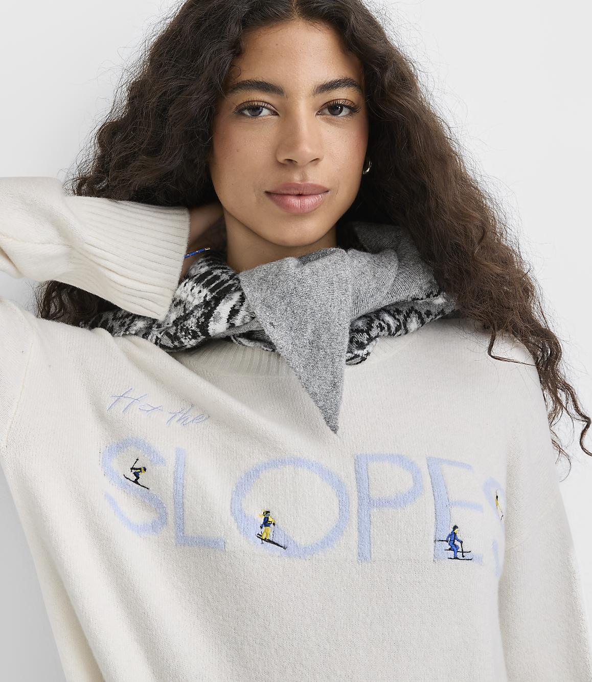 A woman with long curly hair wearing a white graphic sweater and a grey patterned scarf poses against a plain white background.