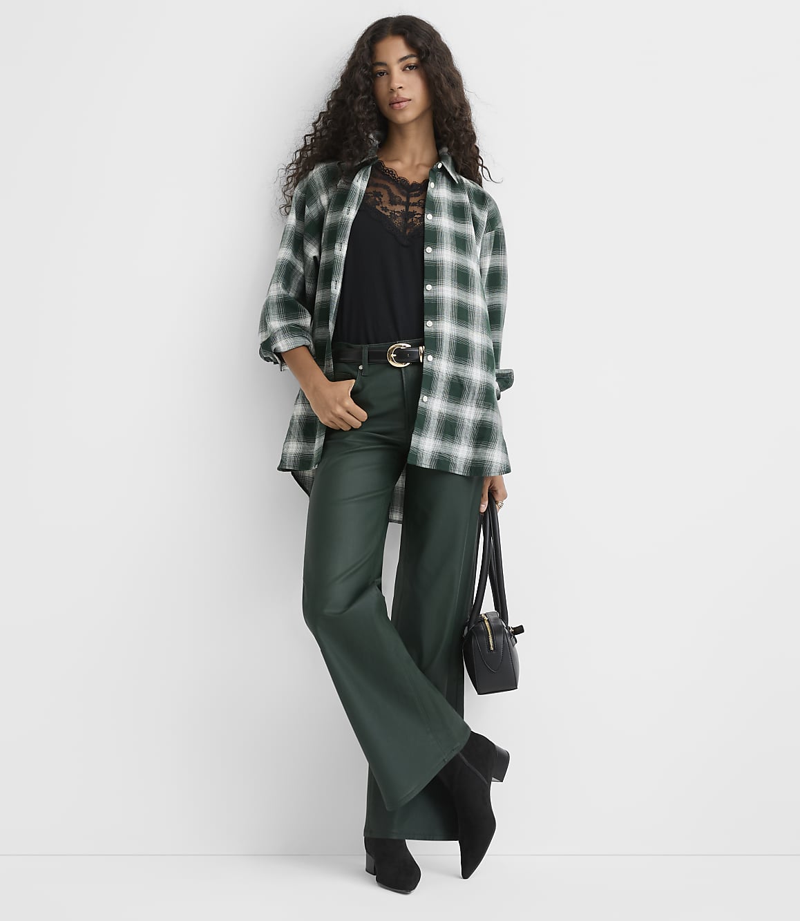A woman wearing a green plaid shirt, black top, and dark green trousers stands against a plain white background.
