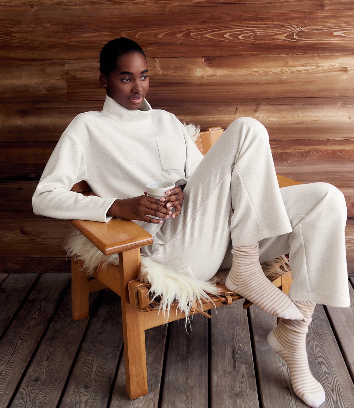 A woman in a white lounge set and striped socks relaxes in a wooden chair against a rustic wood-paneled wall.
