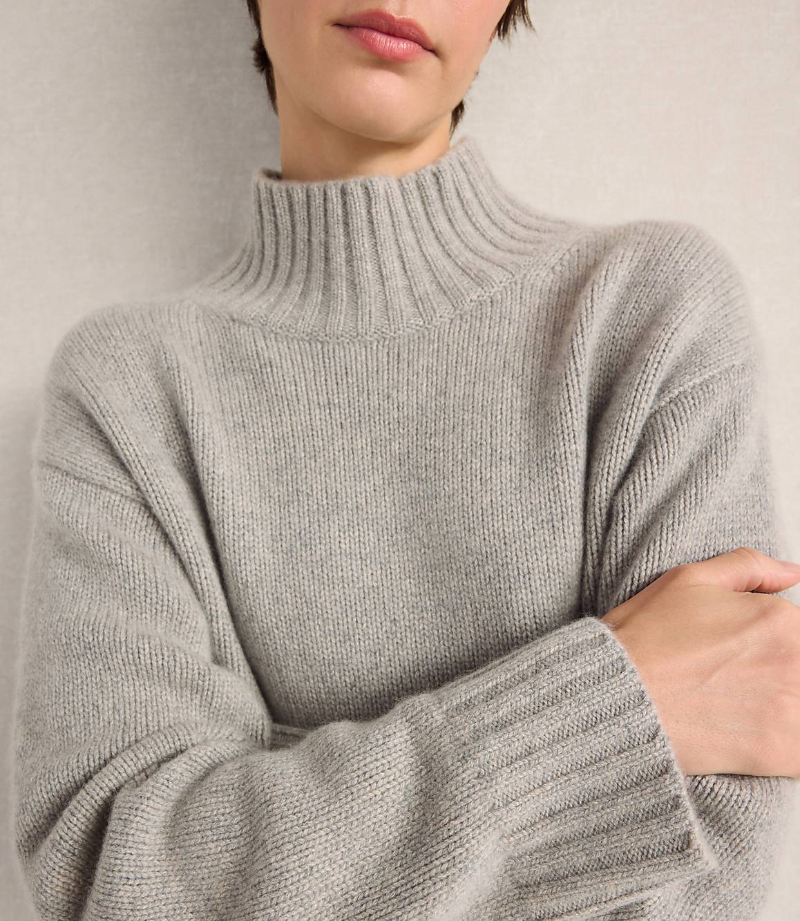 A person wearing a grey ribbed turtleneck sweater stands against a plain, light-colored wall.