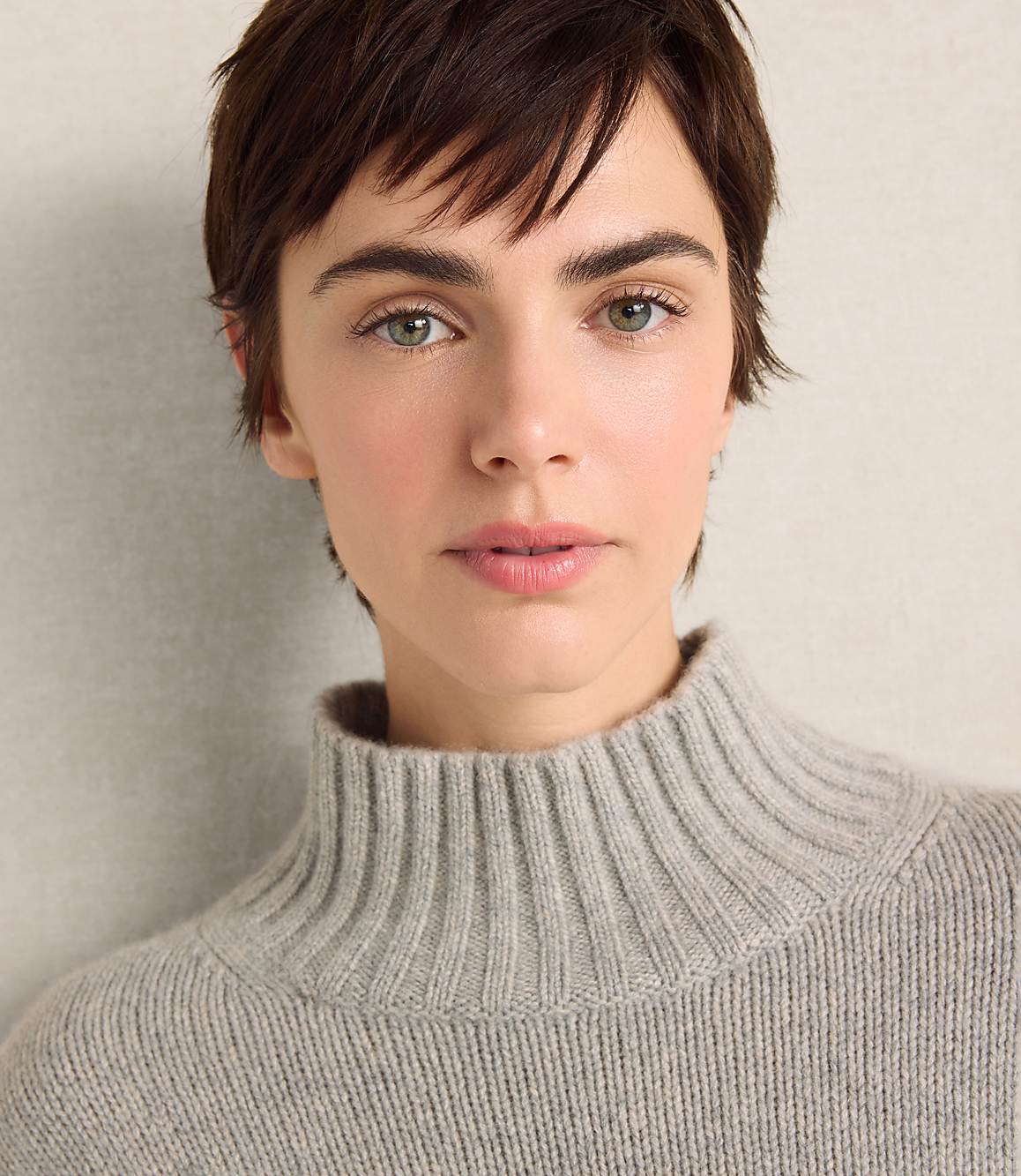 A woman with short brown hair wearing a grey turtleneck sweater poses against a plain, light-colored wall.