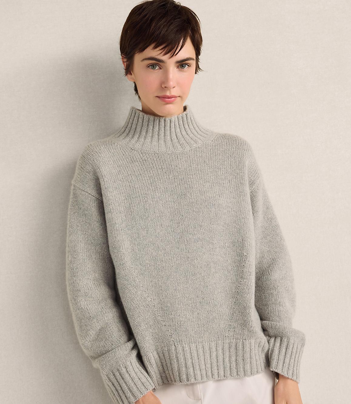 A woman with short brown hair wearing a grey ribbed turtleneck sweater stands against a plain, light-colored wall.