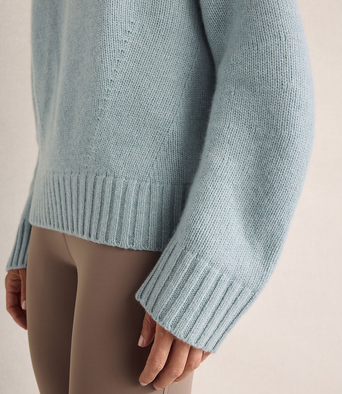 A person wears a light blue knit sweater and brown leggings against a plain, neutral background.