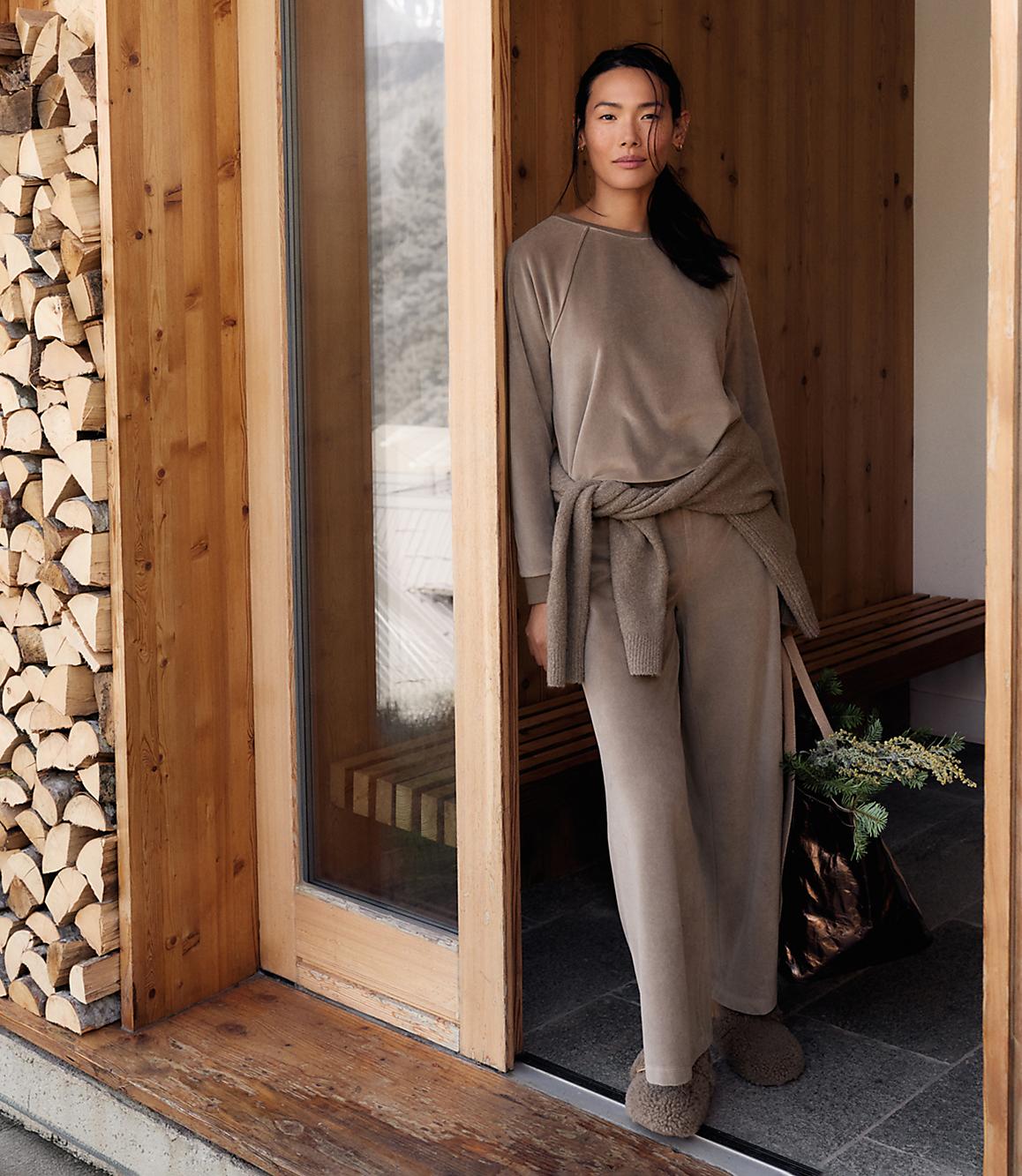 A woman in a taupe lounge set stands in a wooden doorway, with a stack of firewood to her left and a rustic interior behind her.