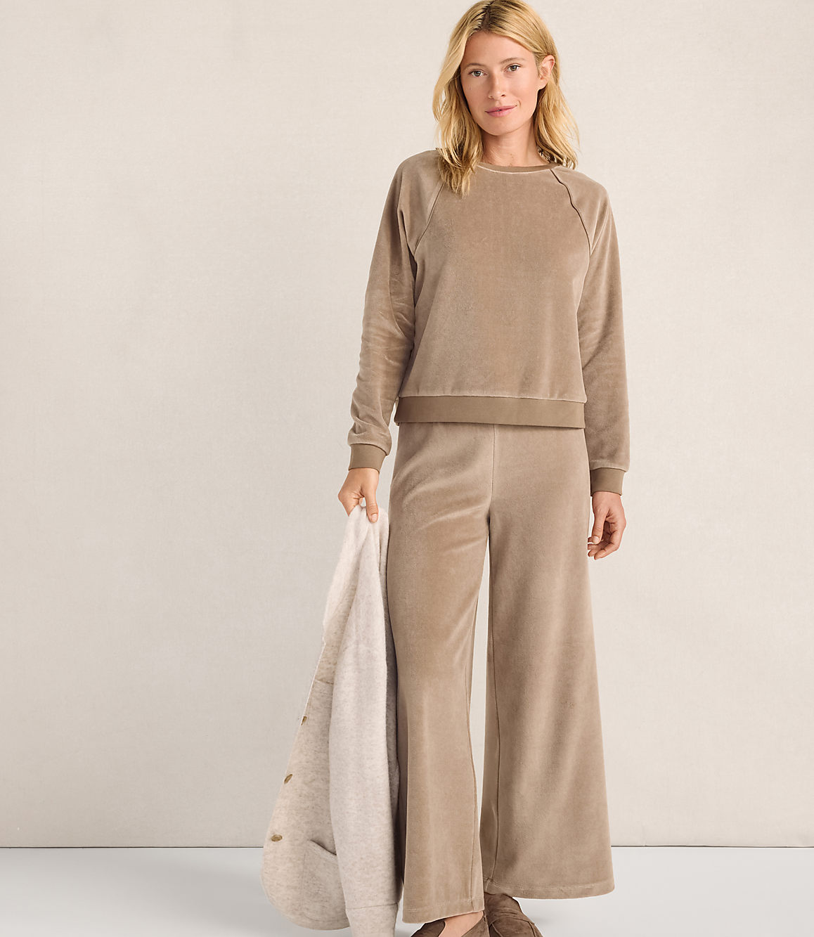 A woman wearing a tan velvet lounge set stands against a plain, light-colored wall while holding a cream-colored blanket.