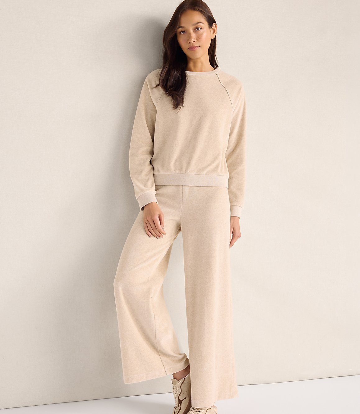 A woman wearing a beige matching sweatshirt and wide-leg pants stands against a plain, light-colored wall.