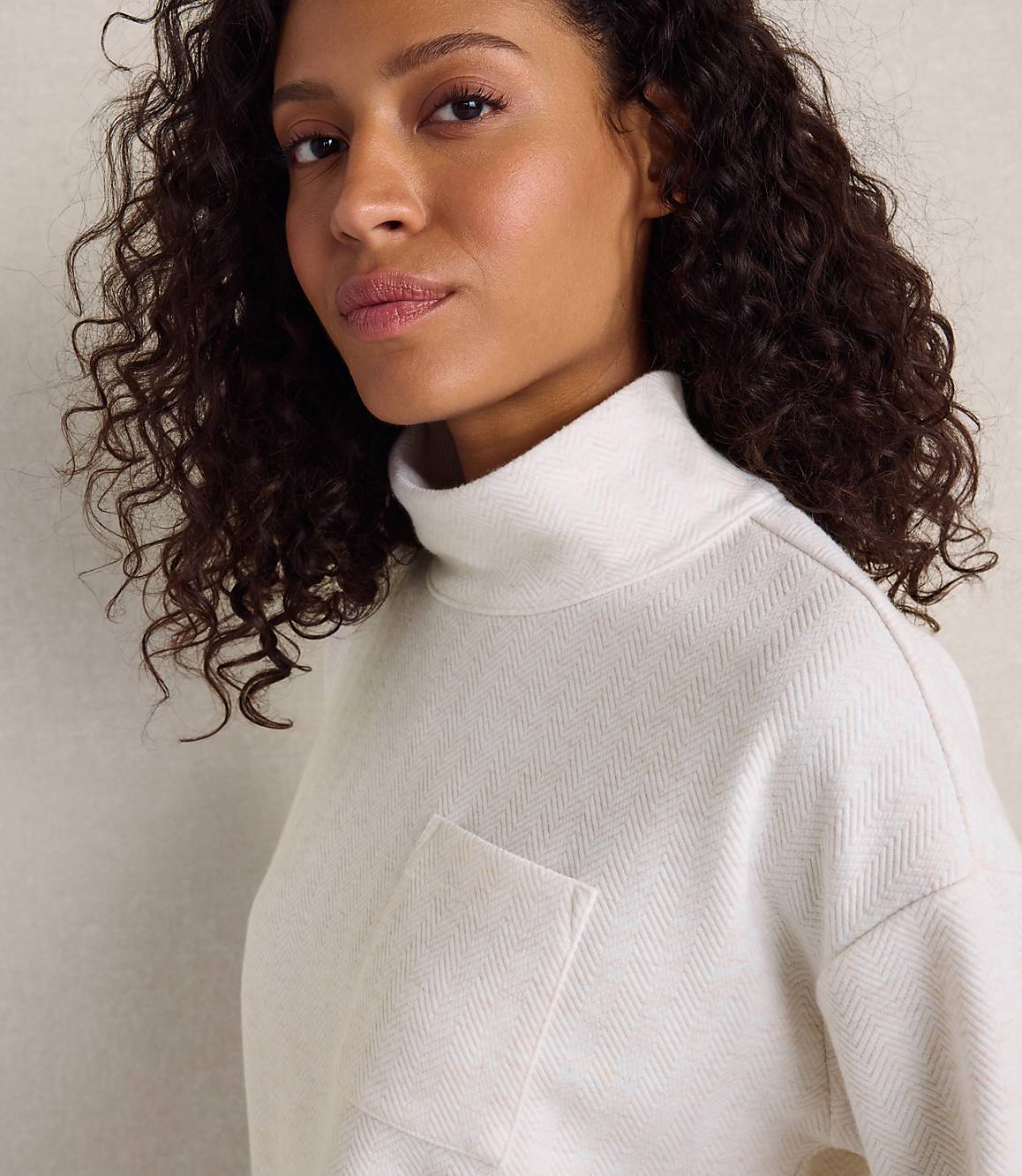 A woman with curly dark hair wearing a white textured turtleneck sweater stands against a plain, light-colored wall.