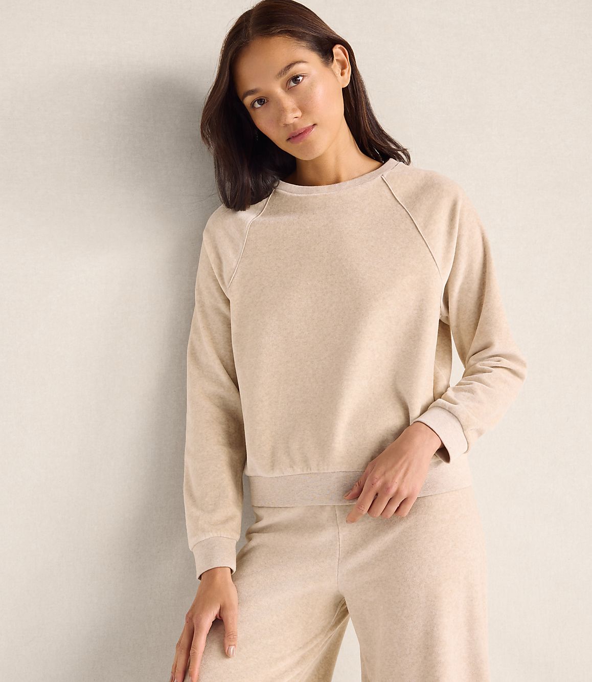 A woman wearing a beige lounge set stands against a plain, neutral-colored wall.
