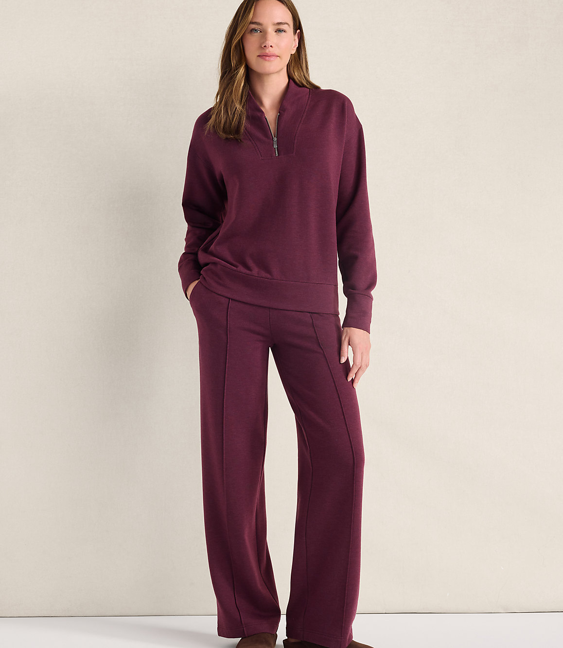 A woman wearing a maroon quarter-zip pullover and matching wide-leg pants stands against a plain, light-colored wall.