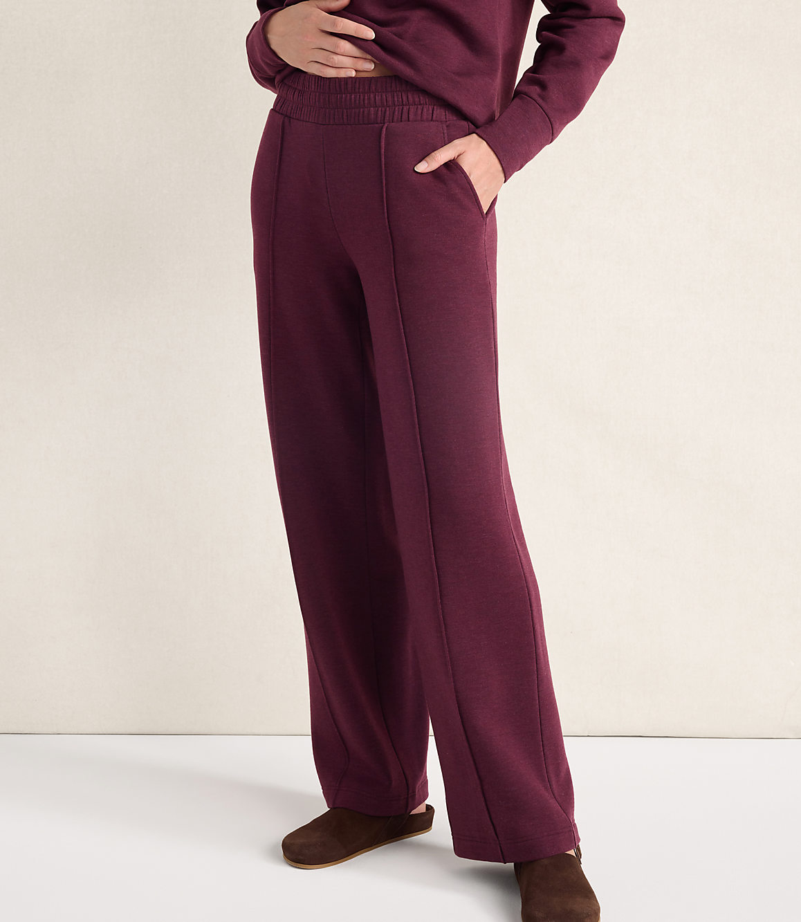 A person wearing a matching maroon sweatshirt and wide-leg trousers stands against a plain, light-colored wall.