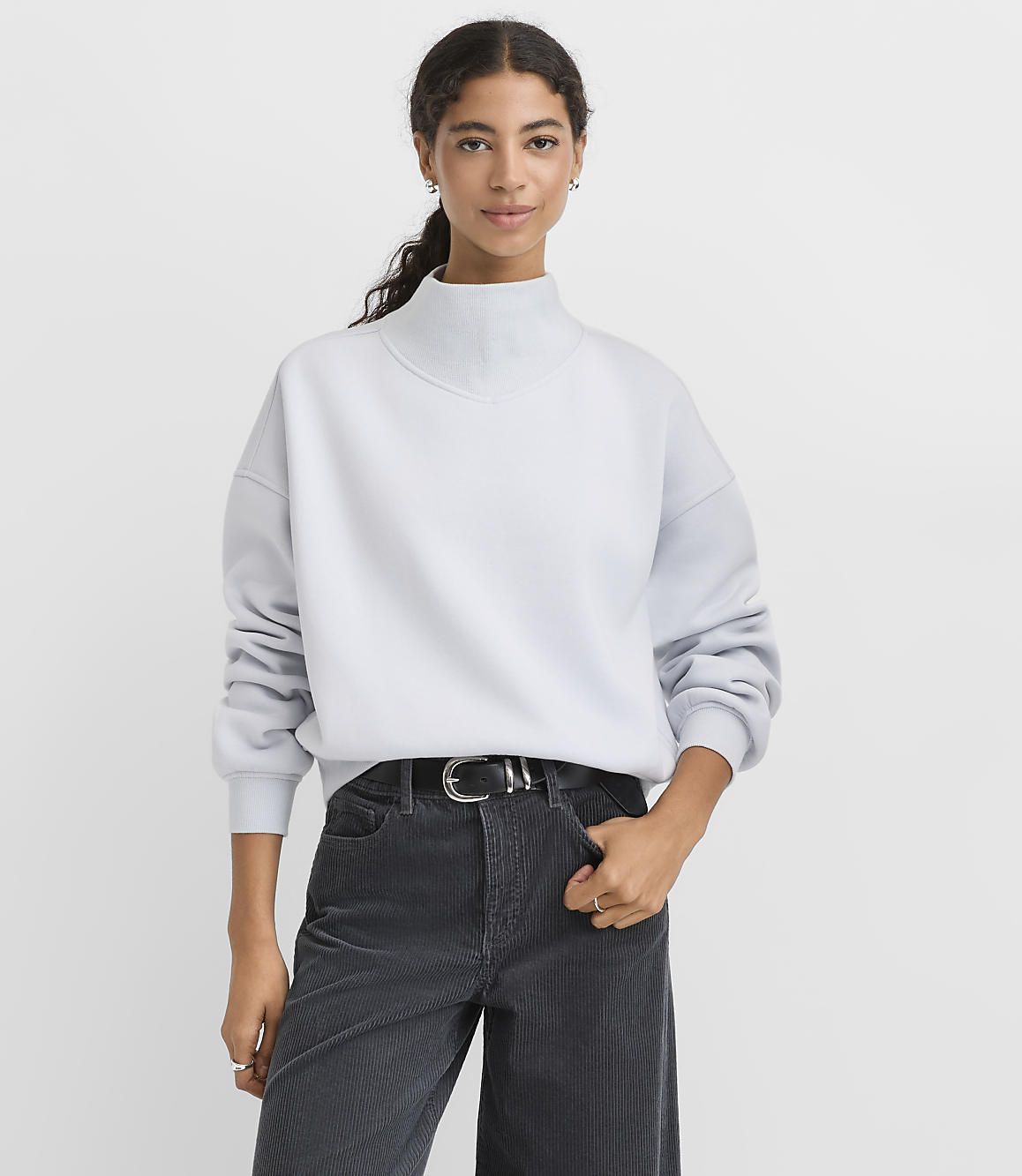 A woman wearing a light blue mock-neck sweatshirt and dark corduroy pants stands against a plain white background.