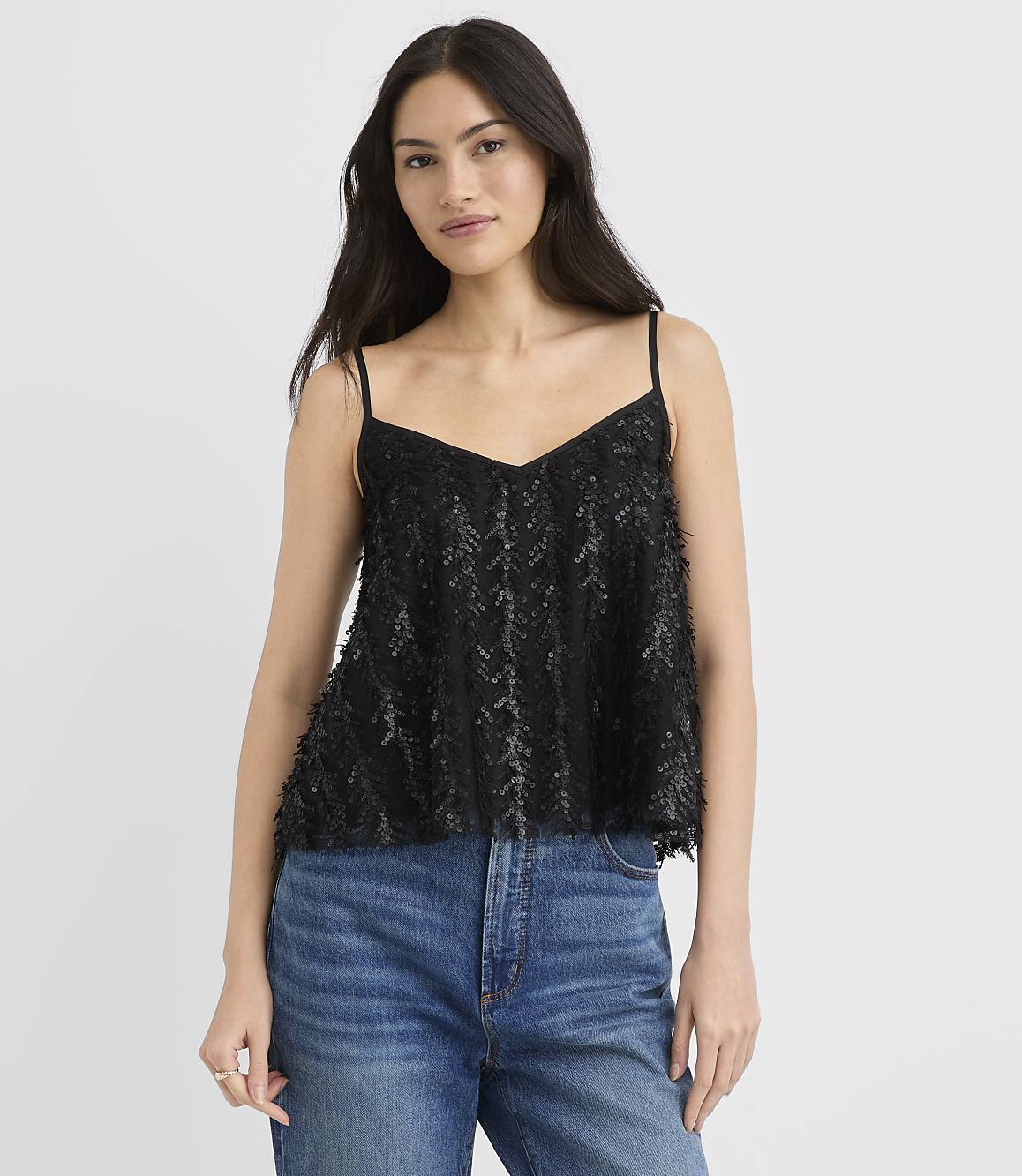 A woman wearing a black sequined camisole and blue jeans stands against a plain white background.