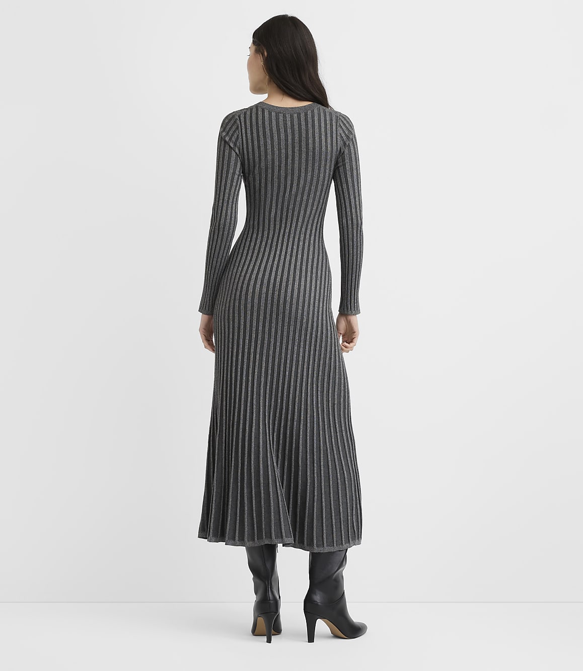 A woman wearing a long-sleeved, ribbed grey maxi dress and black heeled boots stands with her back to the camera against a plain white background.