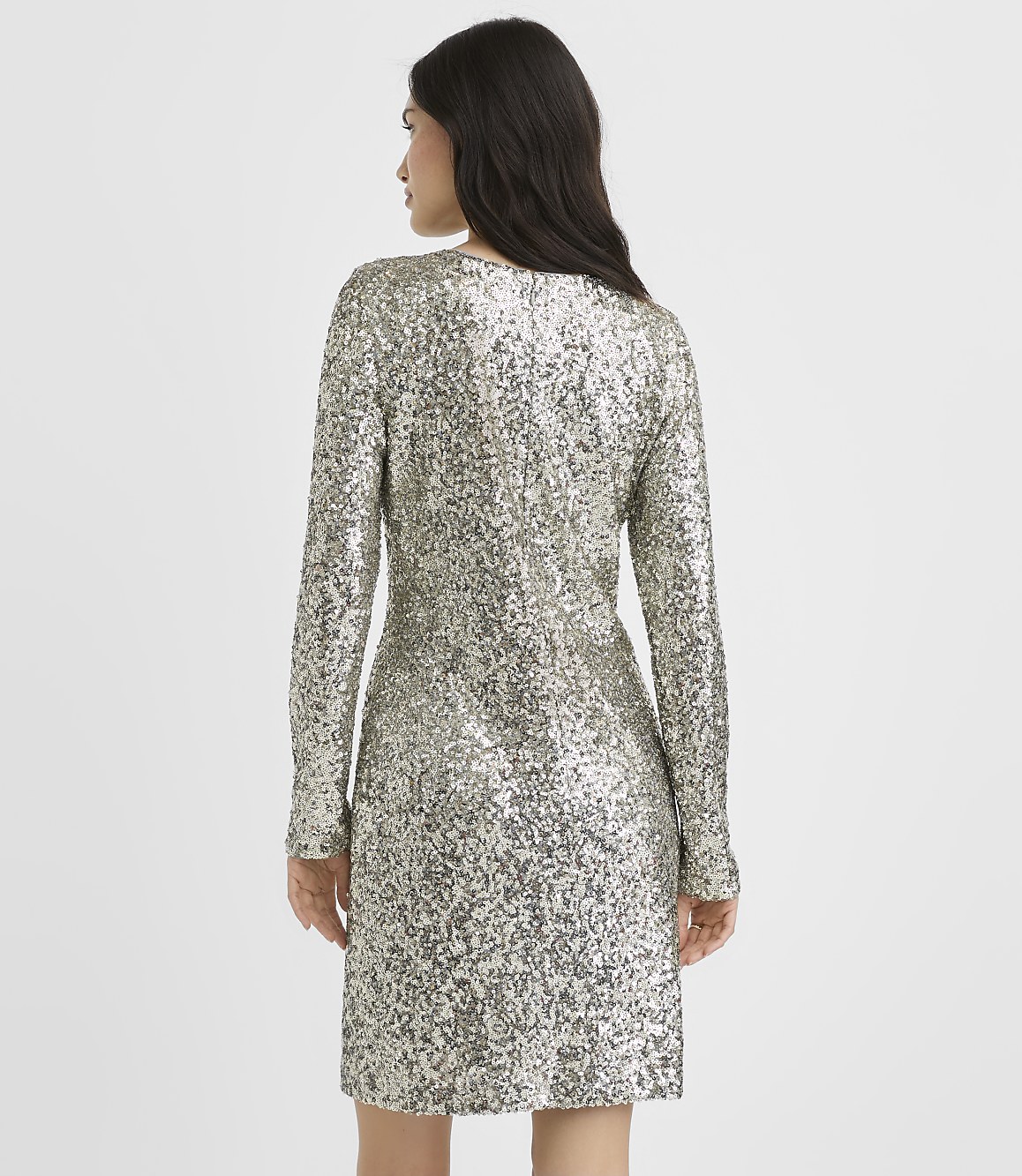 A woman wearing a long-sleeved silver sequin dress stands with her back to the camera against a plain white background.