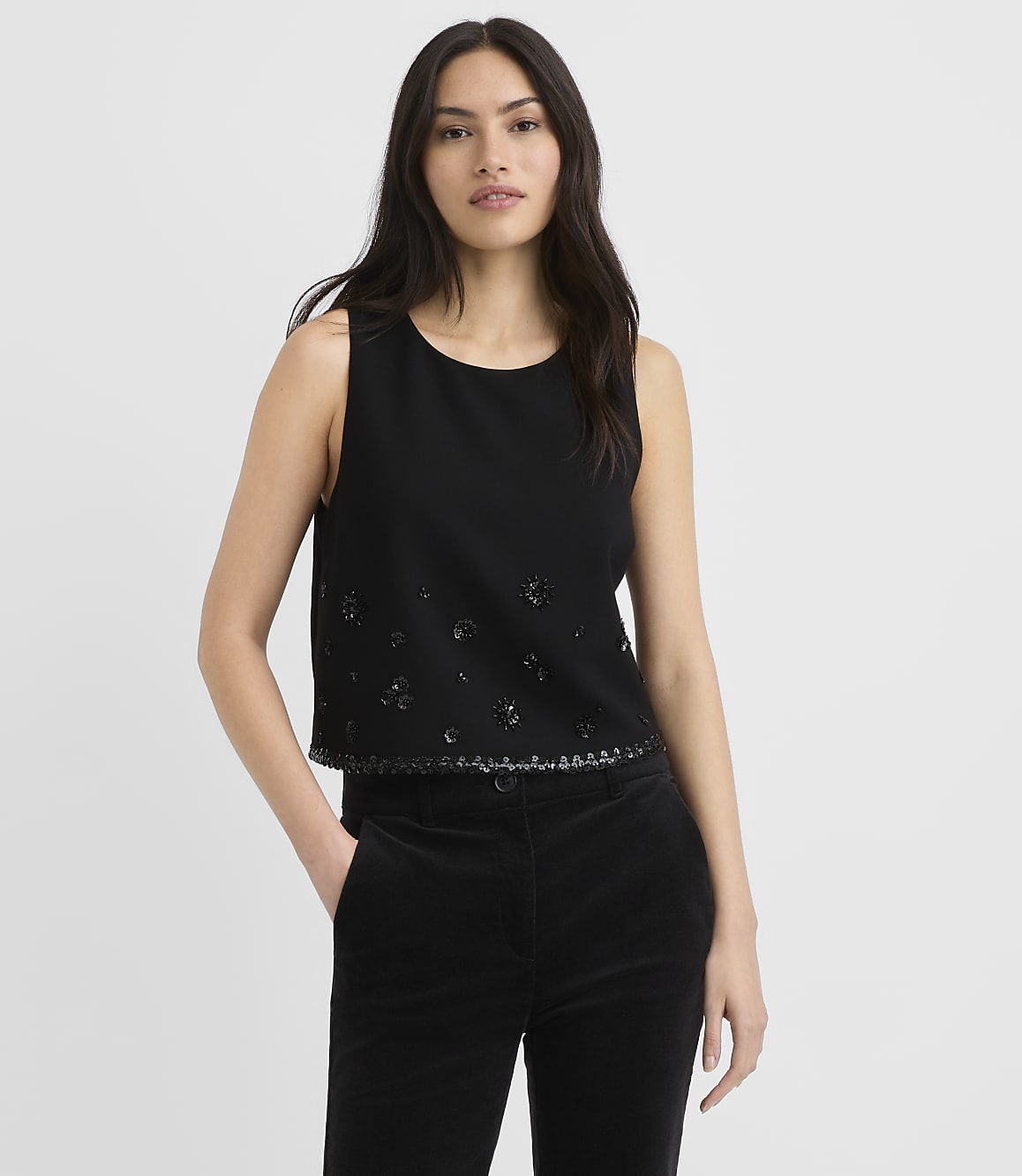 A woman wearing a black sleeveless top and black trousers stands against a plain white background.