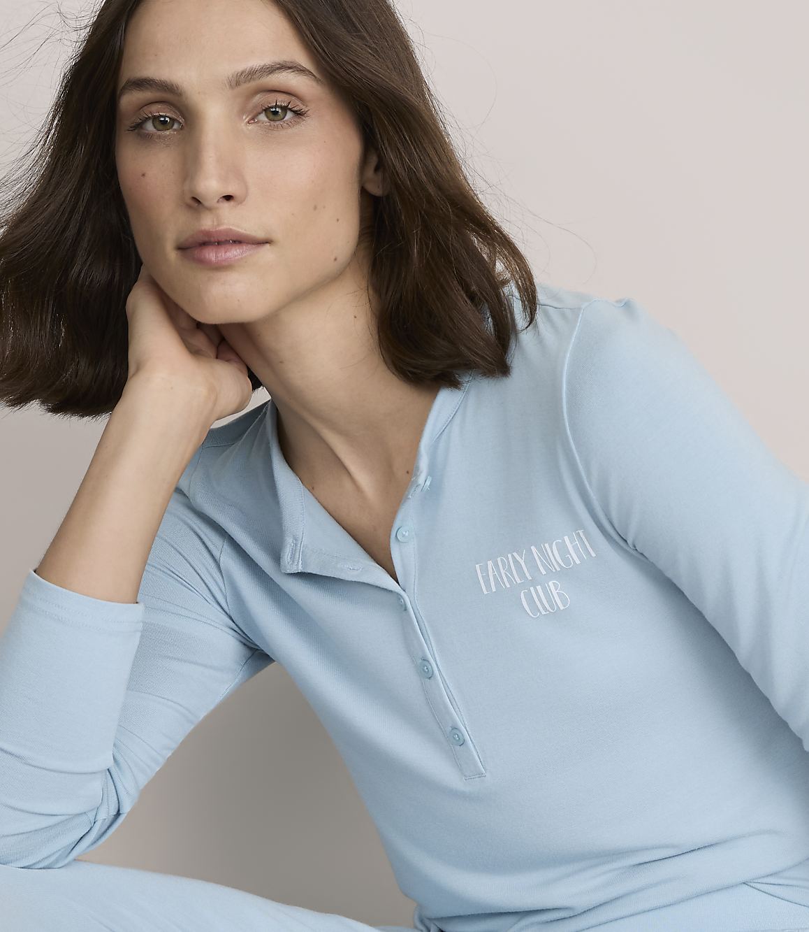 A woman with brown hair wearing light blue pajamas rests her chin on her hand against a plain, neutral background.