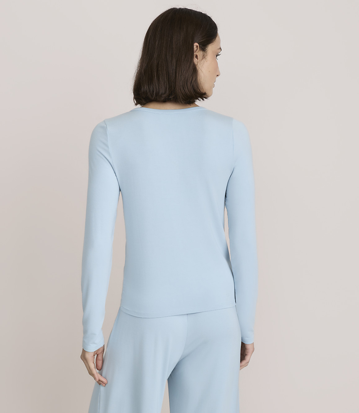 A woman wearing a light blue long-sleeved top and matching pants stands with her back to the camera against a plain, neutral background.