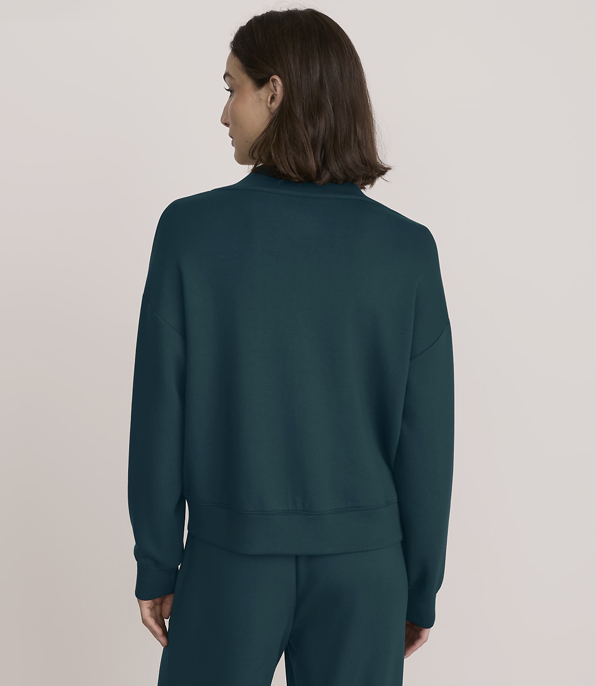 A woman wearing a dark teal sweatshirt and matching pants stands with her back to the camera against a plain, light-colored background.