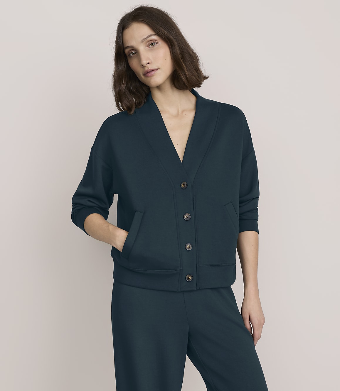 A woman wearing a dark teal button-up cardigan and matching trousers stands against a plain, light beige background.
